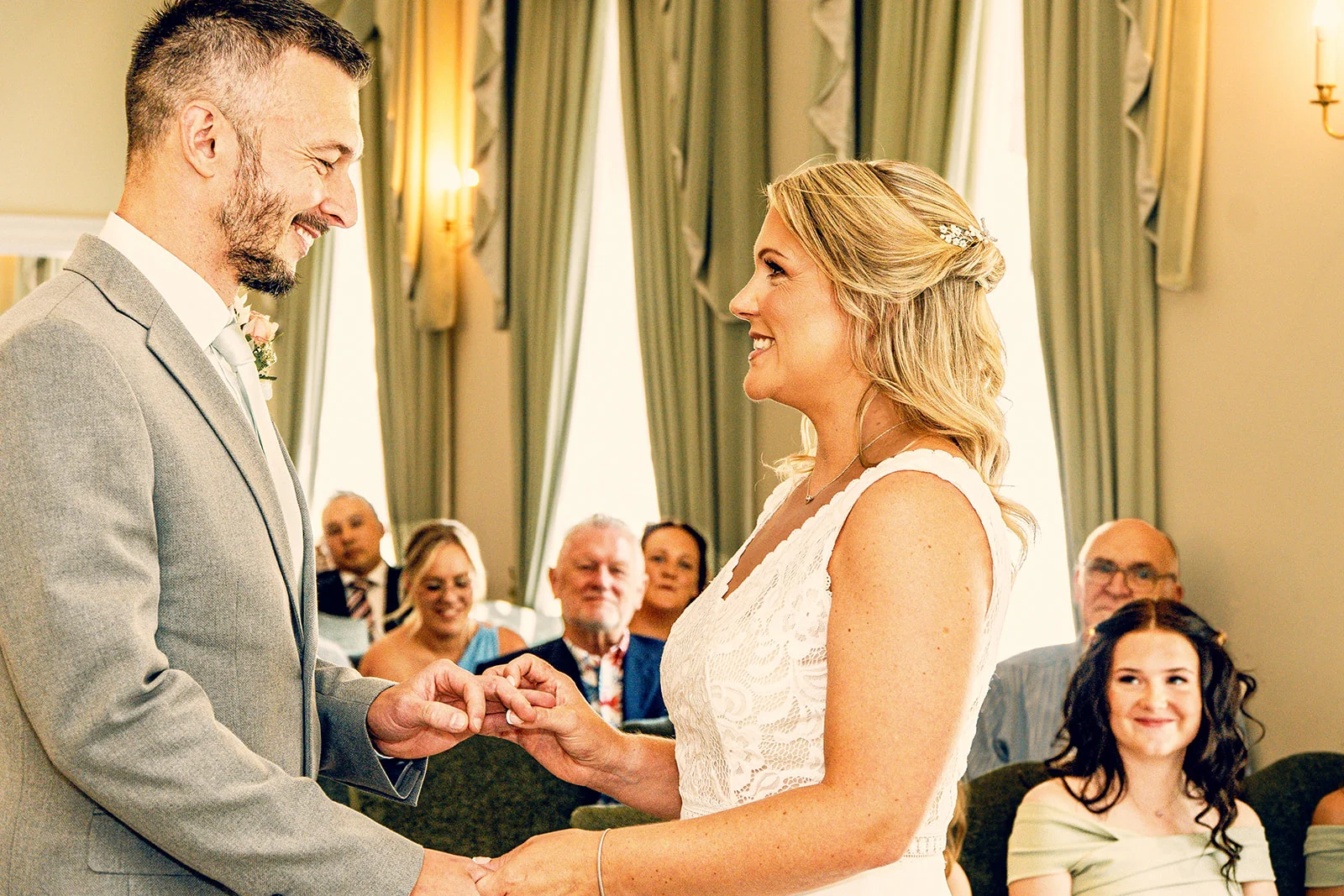 A smiling groom places a ring on the bride's finger during their indoor civil ceremony while seated guests look on.