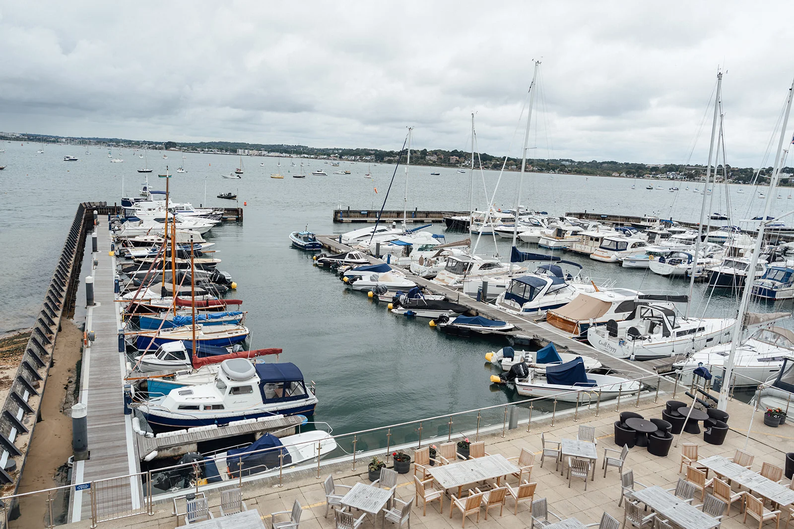 An aerial view of a bustling marina filled with moored boats and yachts under an overcast sky, next to an outdoor seating terrace.