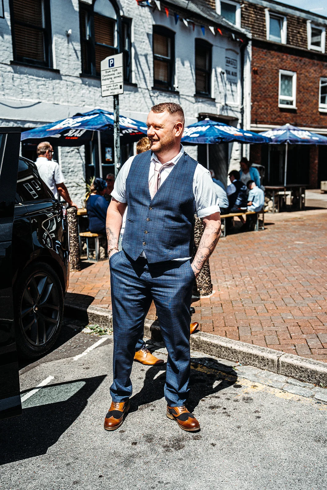 A smiling groom with tattooed arms poses outside a pub on a sunny day wearing a blue checked waistcoat and trousers with brown brogues.