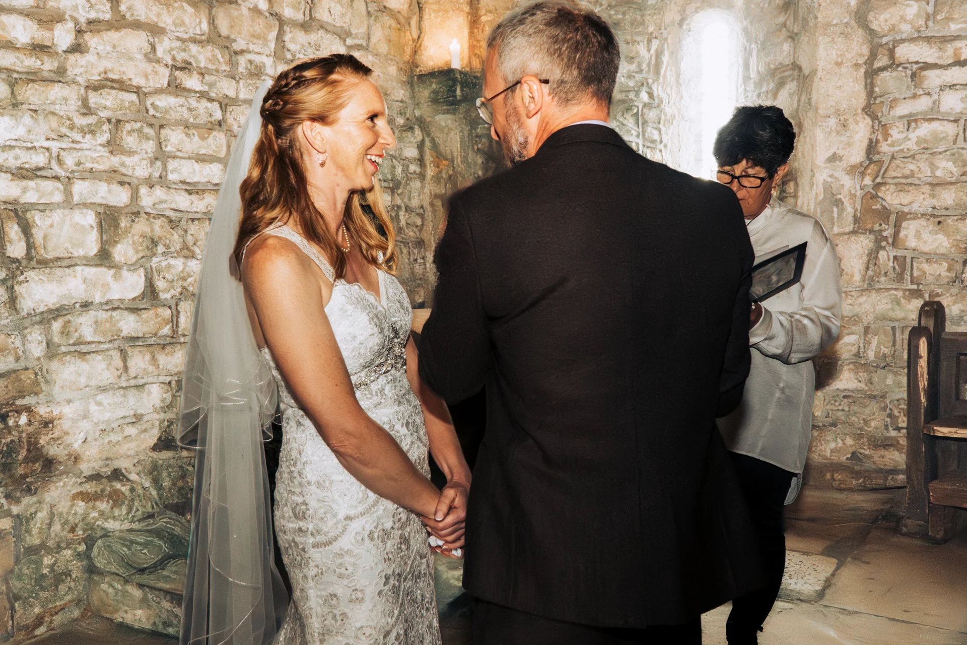 A mature bride and groom smile happily at each other during an intimate indoor ceremony against a rustic exposed stone wall.