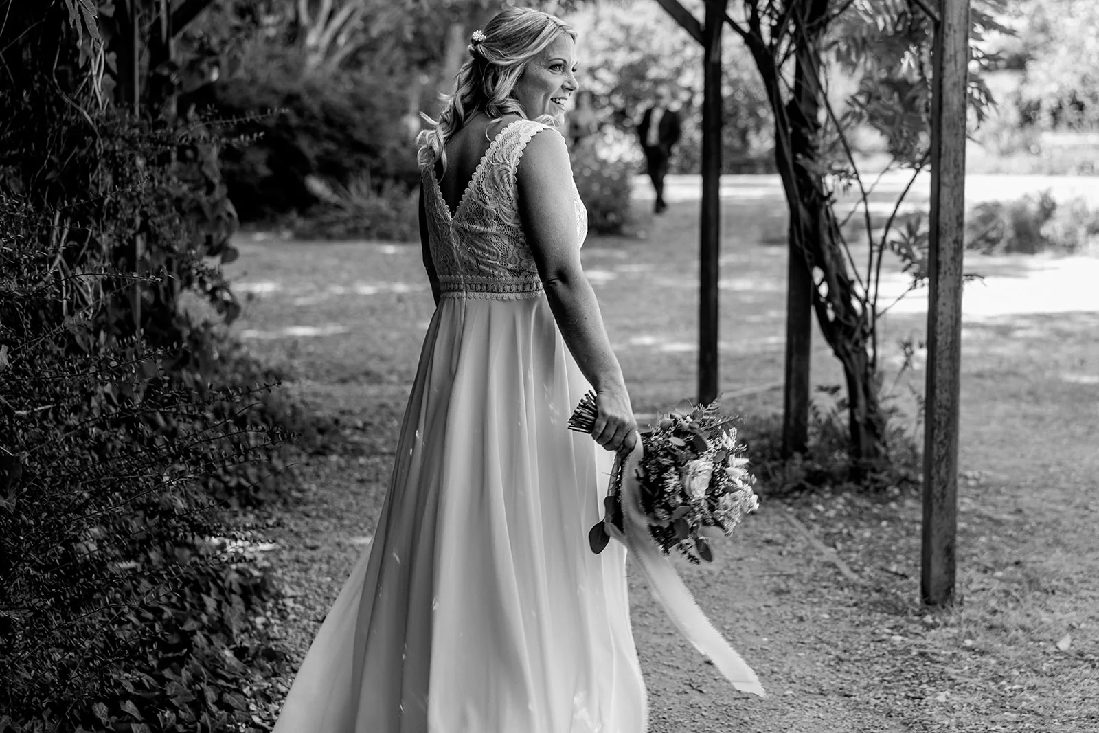 A smiling bride in a lace and chiffon wedding dress looks back over her shoulder while holding a bouquet under a wooden pergola in a sun-dappled garden.