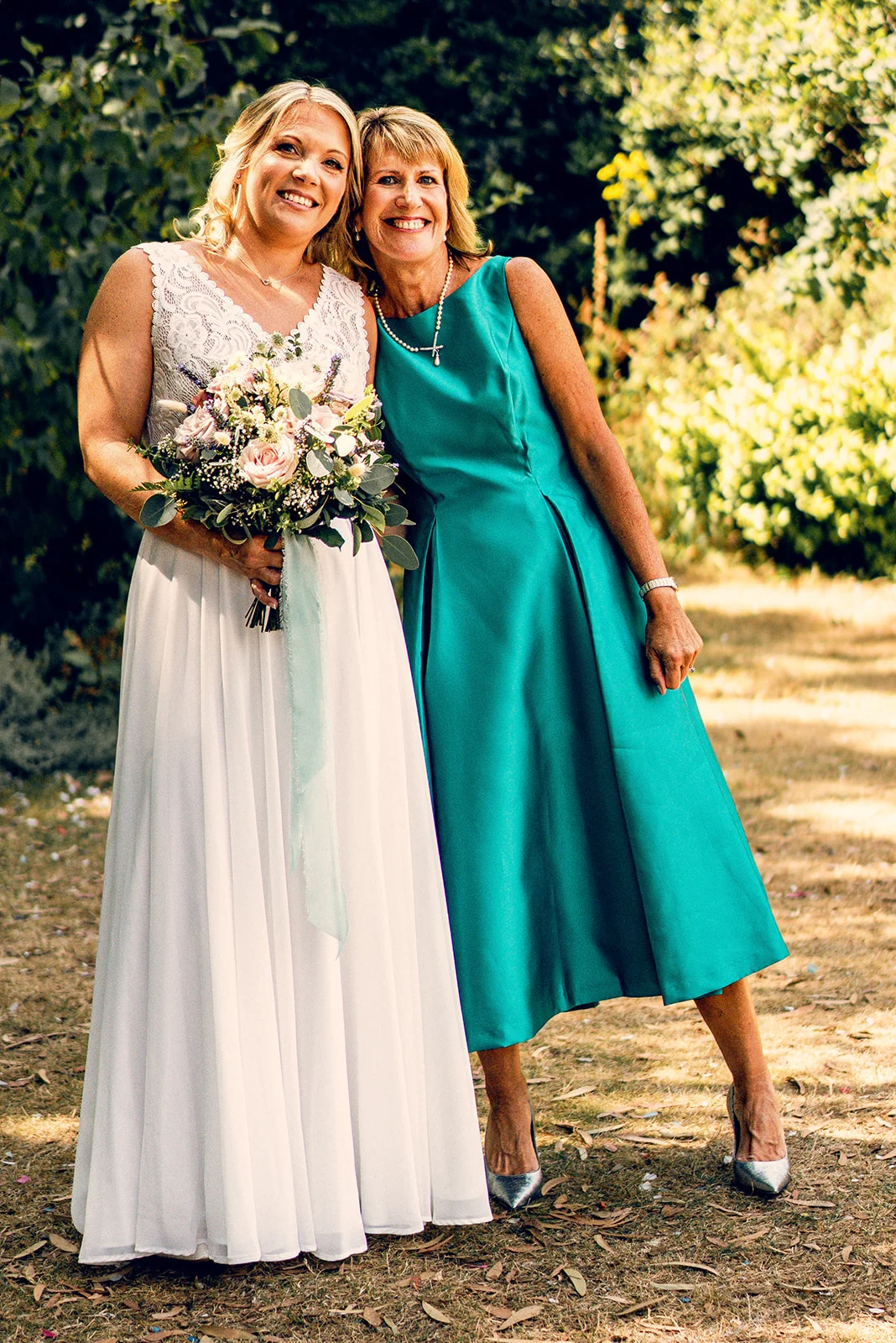 A smiling bride holding a hand tied bouquet stands outdoors alongside the mother of the bride in a vibrant teal dress during the wedding celebration.