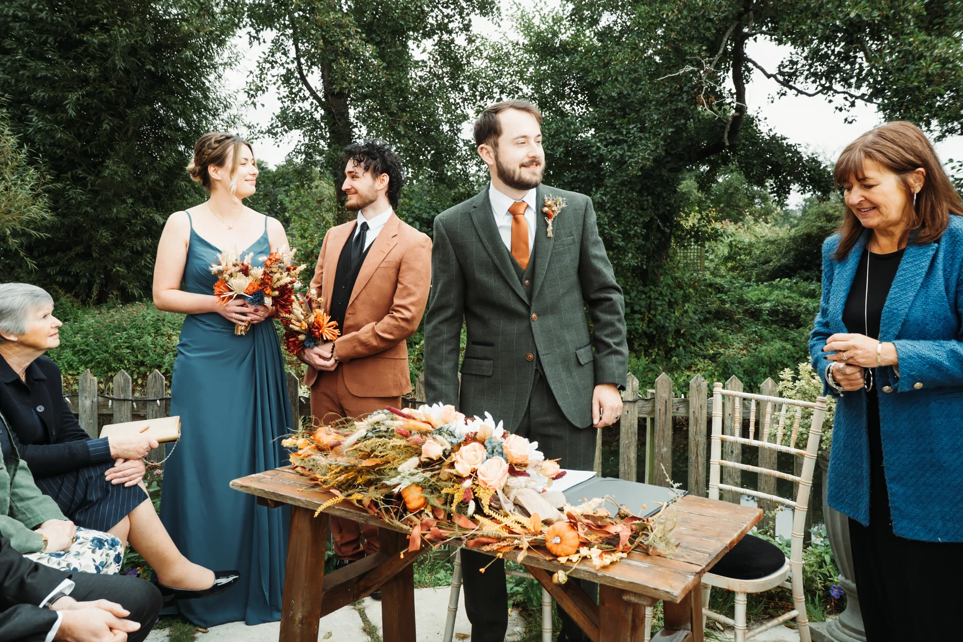 A groom in a green tweed suit and groomsmen stand near an outdoor ceremony table adorned with autumnal foliage, alongside bridesmaids and seated guests.