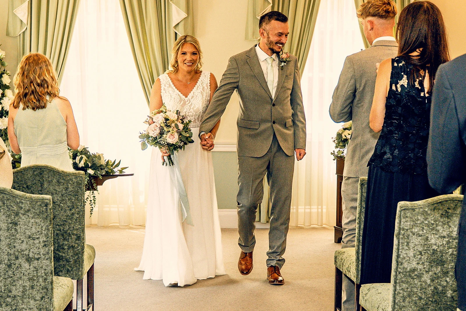 A newlywed couple happily walk hand in hand down the aisle after their ceremony, smiling at guests while the bride holds a delicate floral bouquet.