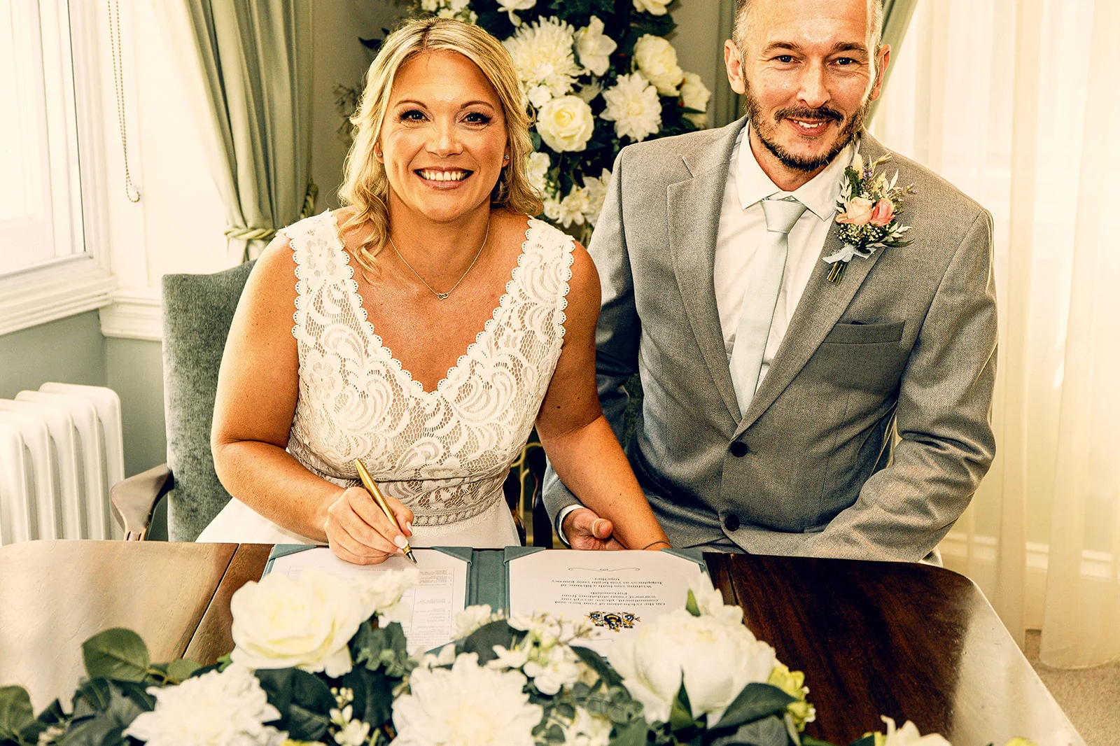A smiling bride and groom sit together at a wooden table while signing the marriage certificate during their civil ceremony.