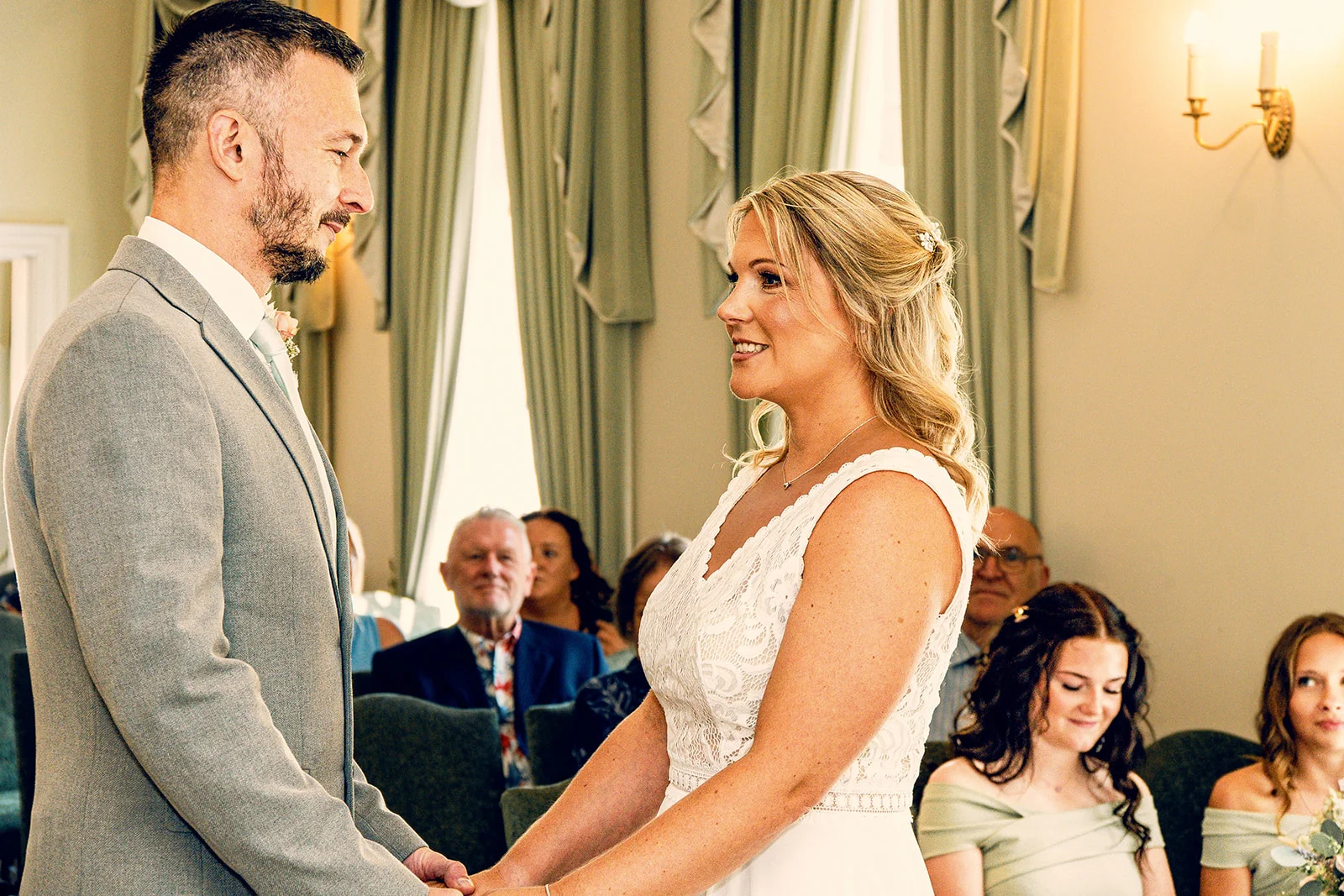A groom in a grey suit and a bride in a lace wedding dress stand face-to-face exchanging vows during an indoor wedding ceremony.
