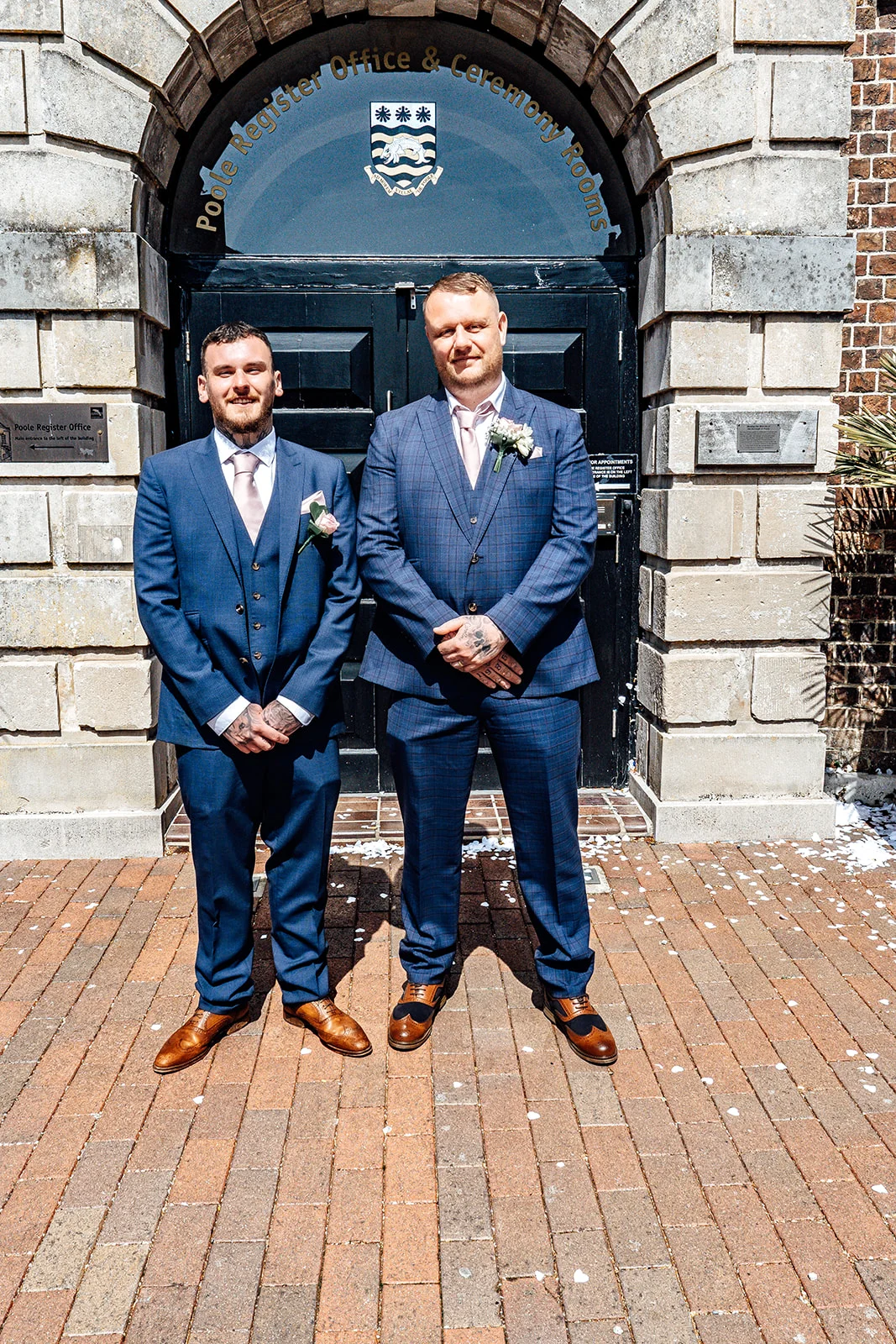 Two grooms stand proudly outside Poole Register Office, wearing matching blue suits with pink ties and buttonholes after their ceremony.