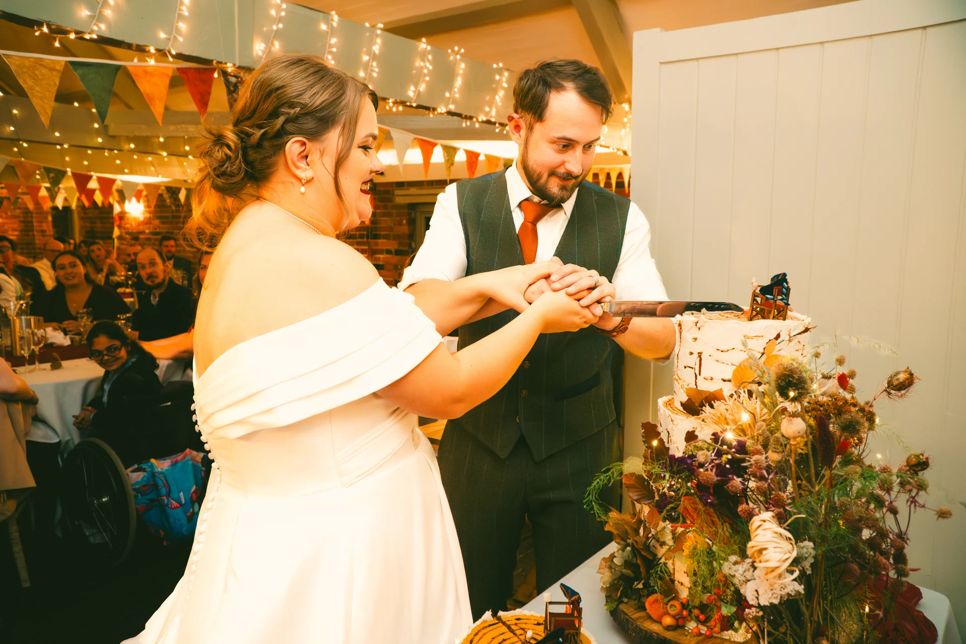 The bride and groom smile together as they cut into a tiered wedding cake adorned with dried florals inside a rustic barn decorated with fairy lights.