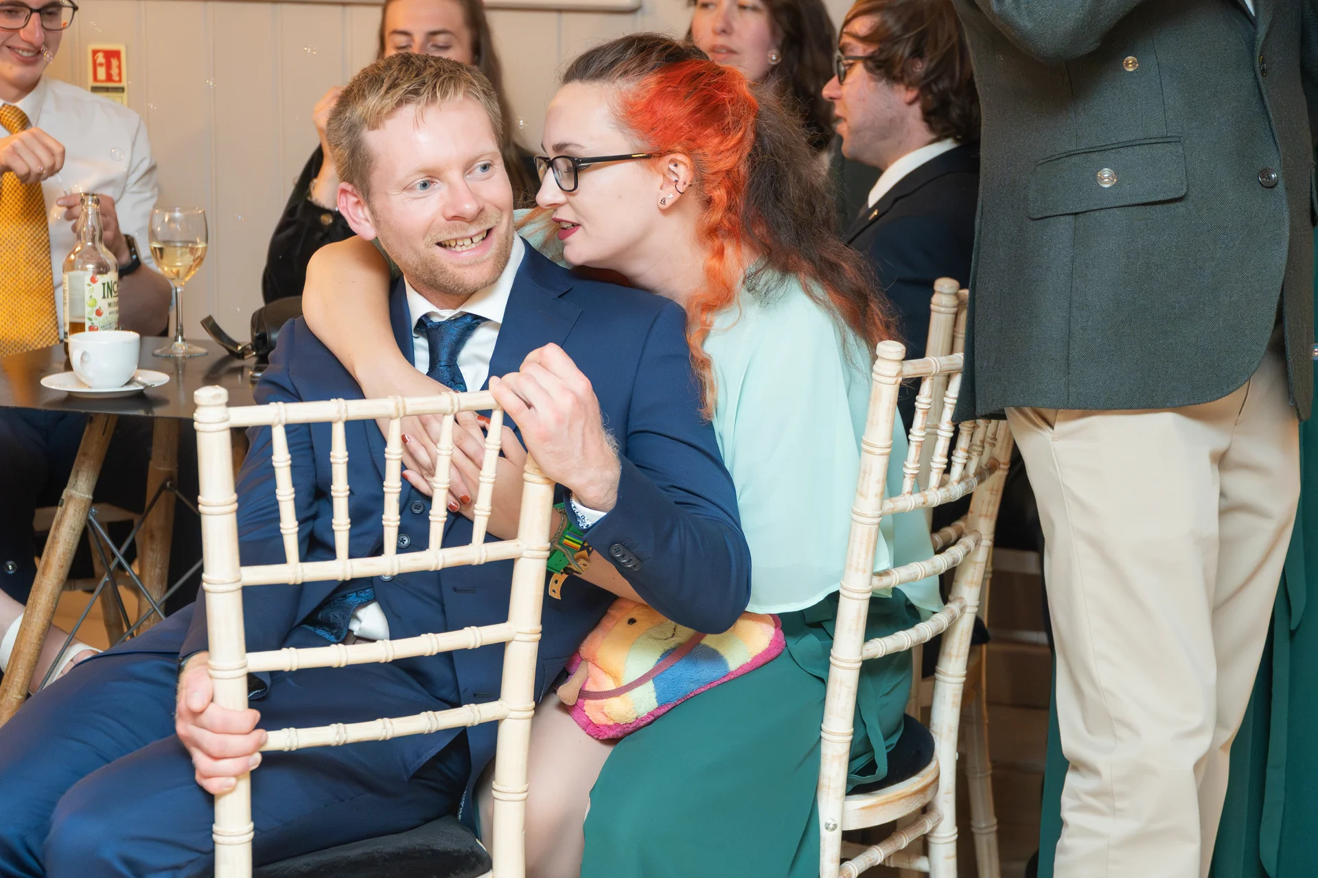 A groom in a navy suit sits with a guest having bright red and dark hair while guests mingle around them during the wedding reception.