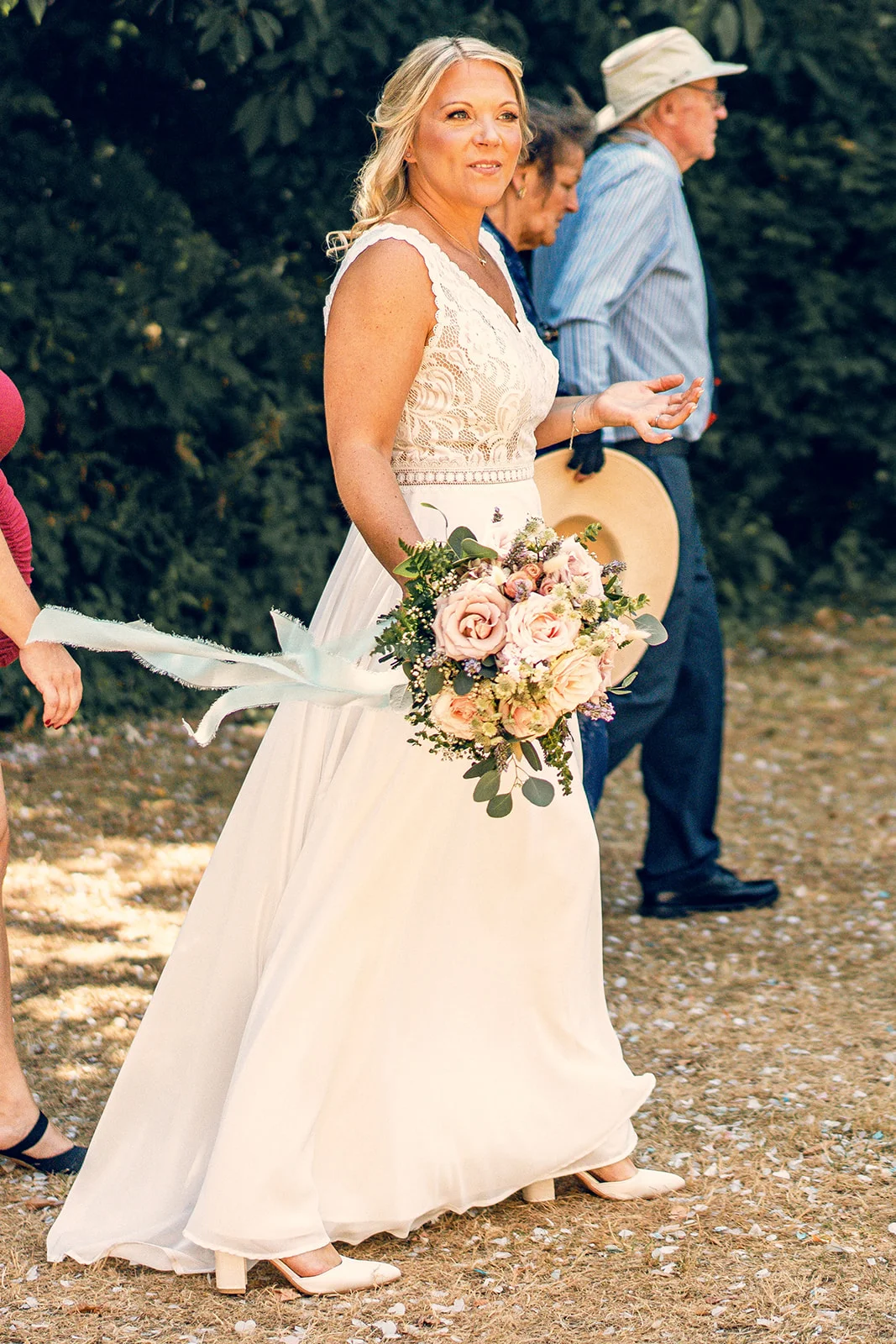 A blonde bride in a lace top and flowing white gown walks outdoors, holding a hand-tied bouquet with trailing ribbons under dappled sunlight.