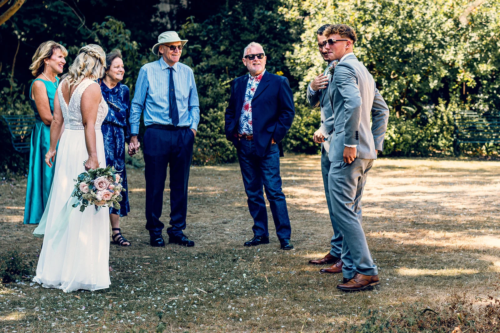 A bride holding a floral bouquet stands outdoors in a sunlit garden, chatting and laughing with guests dressed in smart-casual attire and a grey suit.