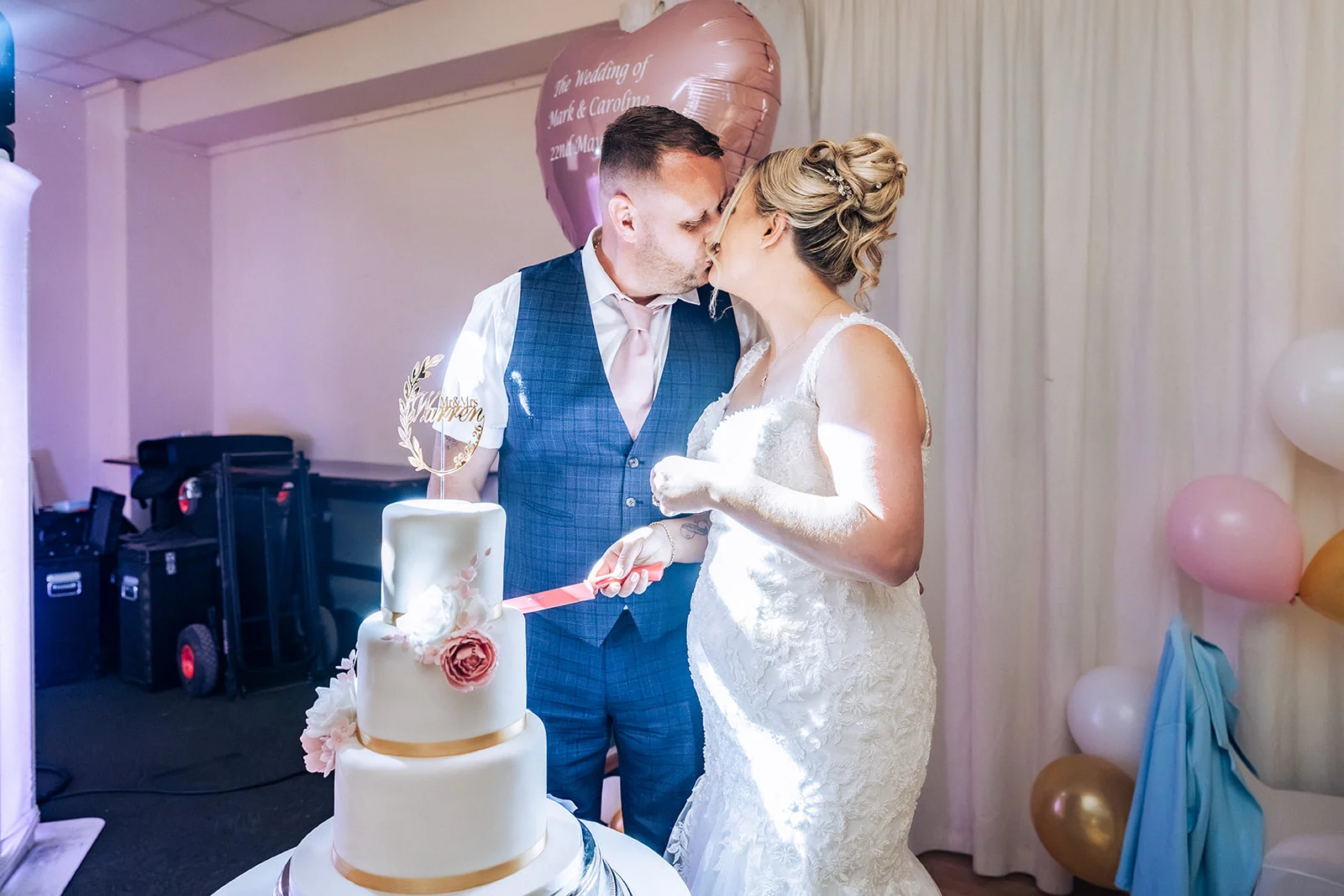 A newlywed couple share a tender kiss while cutting a three-tiered white wedding cake adorned with pink floral details and a custom topper.