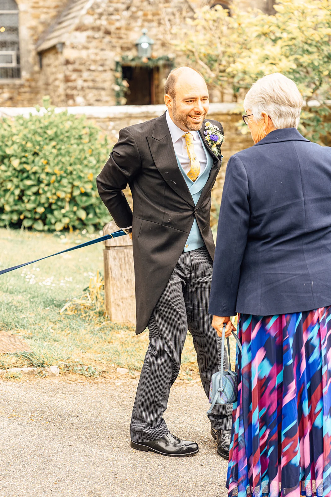 A smiling groom wearing a traditional morning suit with a blue waistcoat and yellow tie chats with a wedding guest outside a stone church.