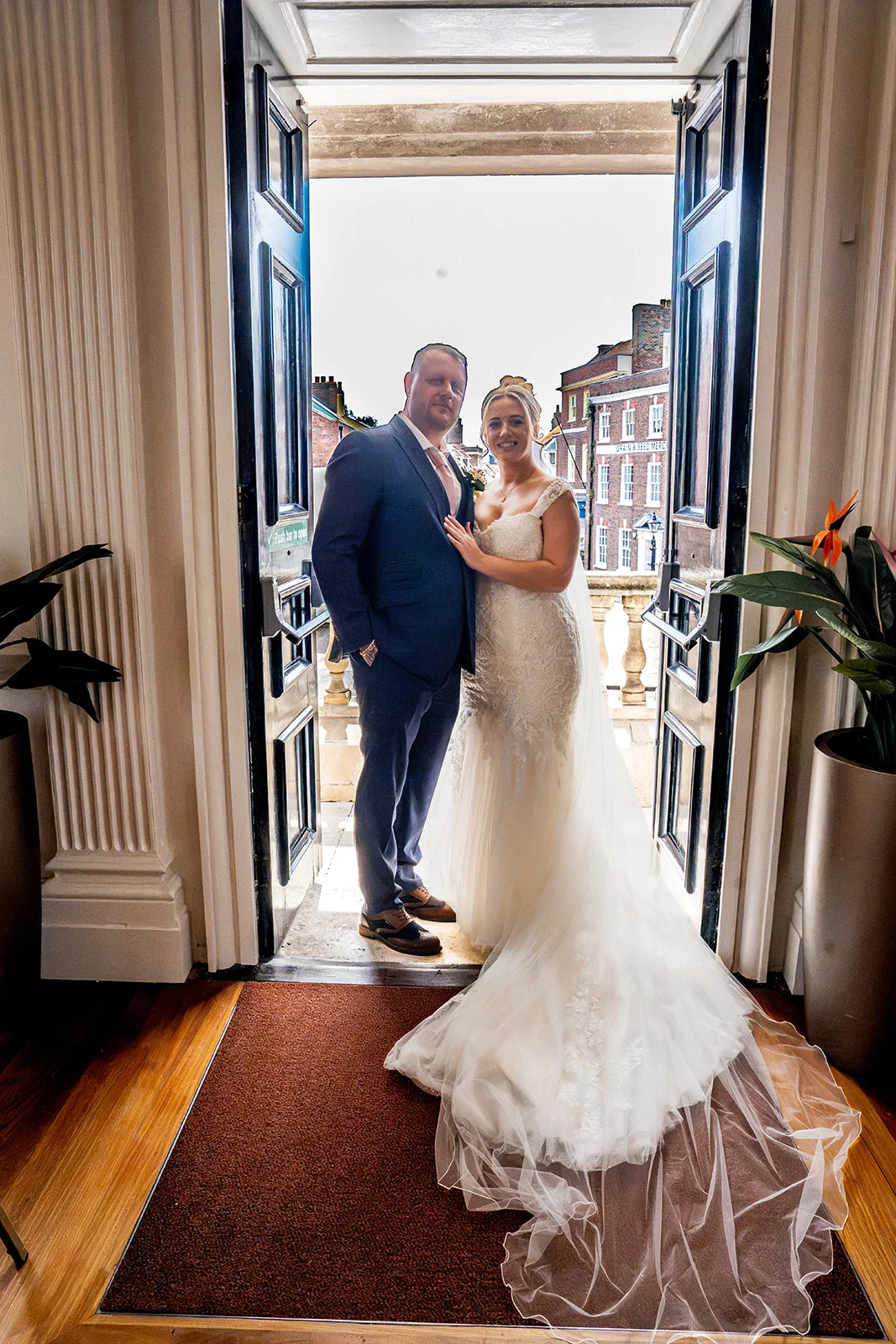 A newlywed couple poses together in the open doorway of a historic town hall, with the bride wearing an elegant lace gown and a flowing veil.
