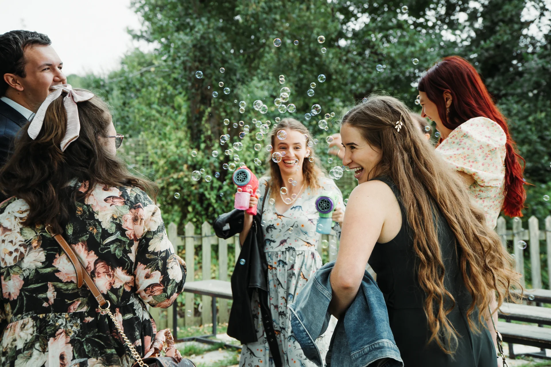 A joyful group of wedding guests laugh and play with bubble blasters in a lush outdoor garden setting during the celebrations.