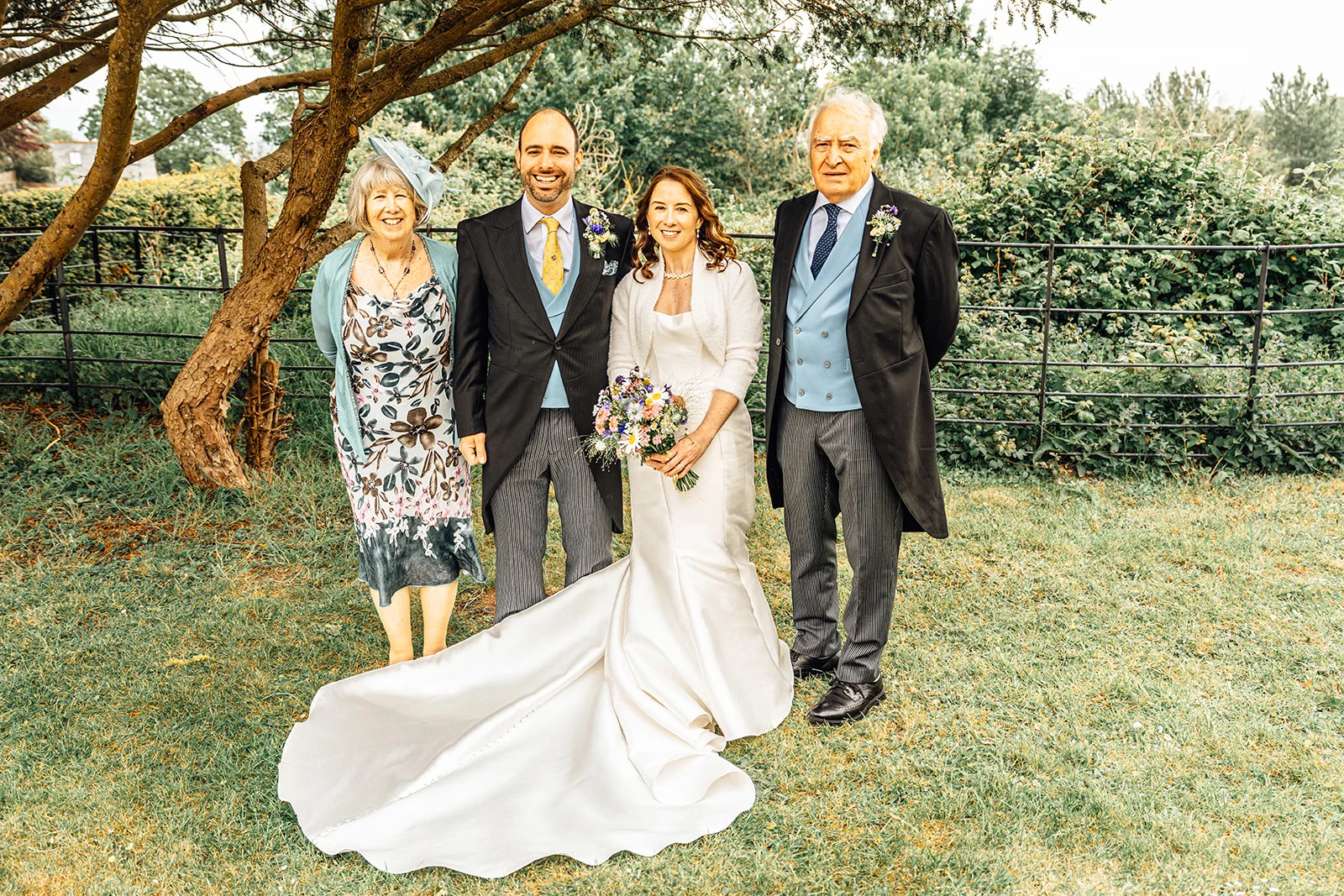 A happy bride and groom pose outdoors with family members on a lawn, flanked by a tree and a metal fence.
