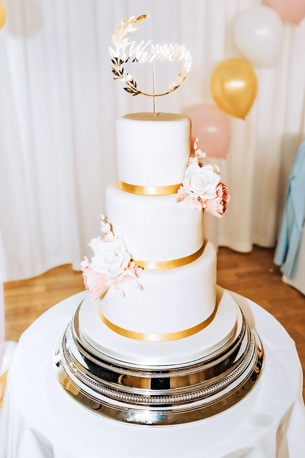 An elegant three-tier white fondant wedding cake adorned with gold ribbon accents and delicate pink and white sugar flowers, displayed on a silver cake stand with balloons in the background.