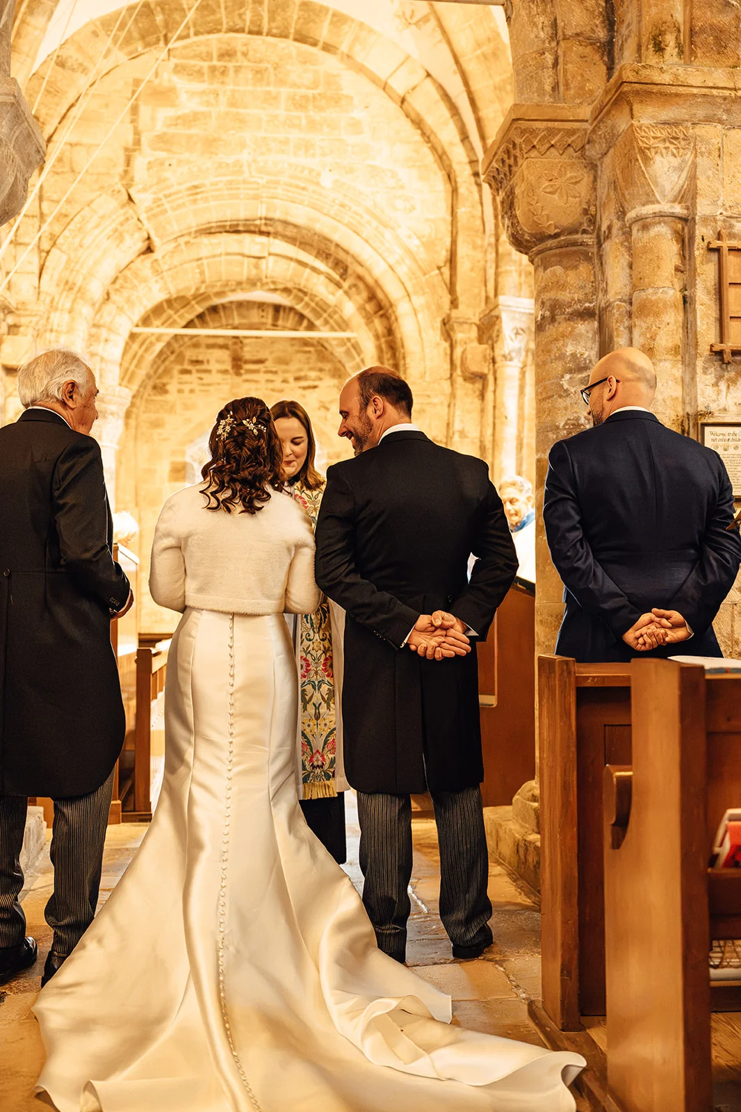 A bride in a satin wedding gown with a faux fur stole and groom in morning dress stand with wedding guests inside a historic stone church during the ceremony.