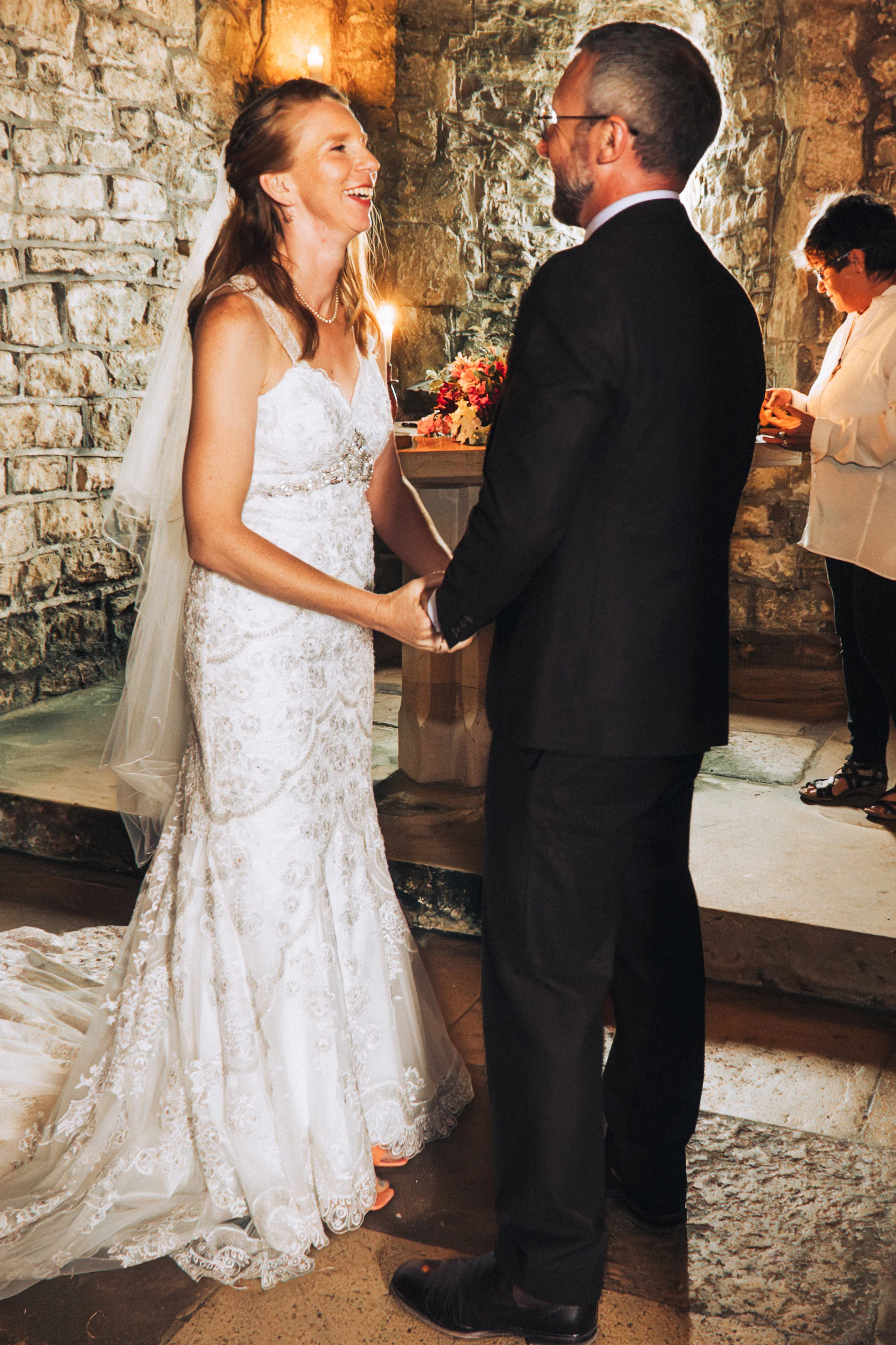A joyful bride and groom hold hands and laugh together during their ceremony inside a rustic stone venue.