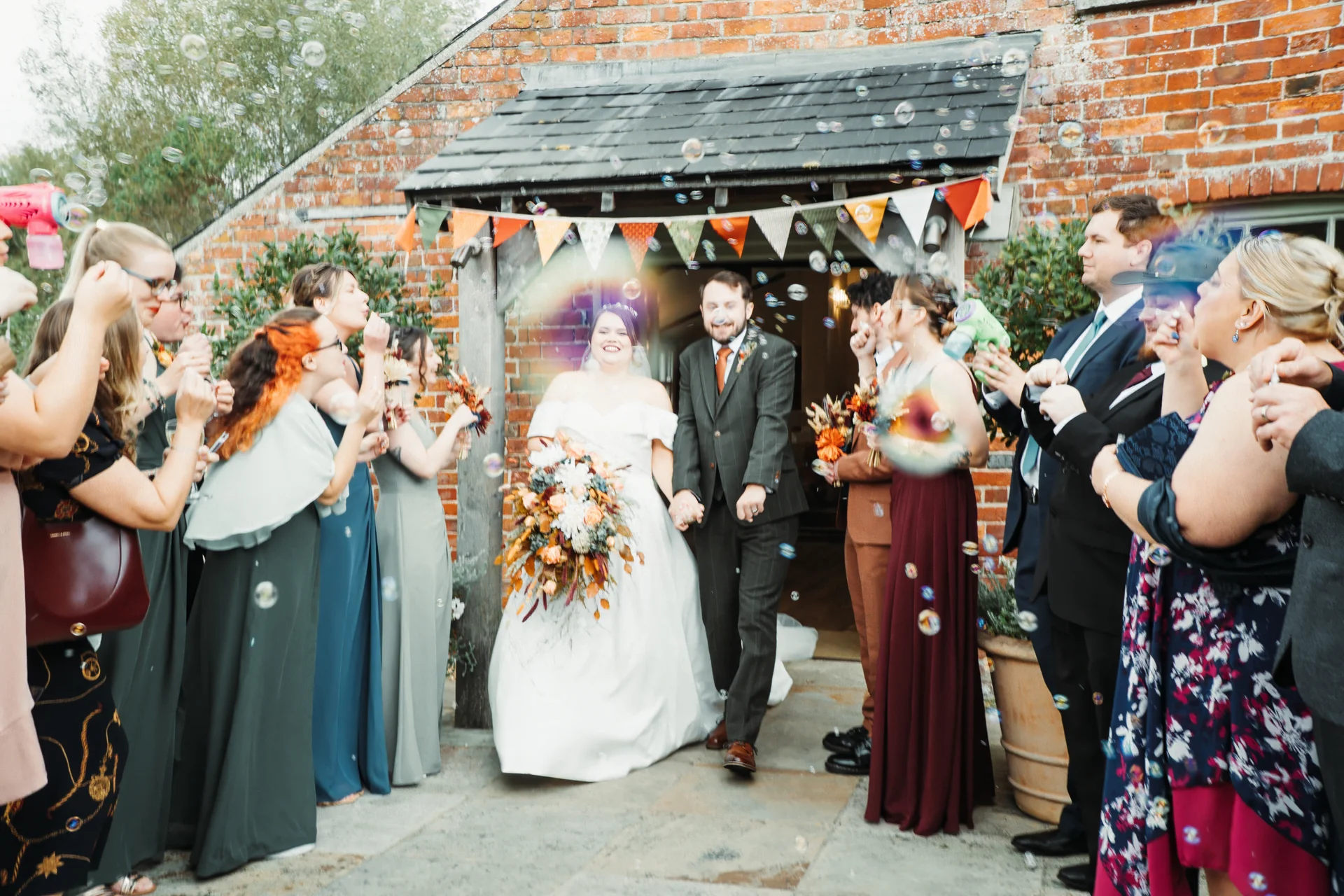 Newlyweds joyfully exit a rustic brick venue holding hands amidst a shower of floating bubbles from cheering guests.