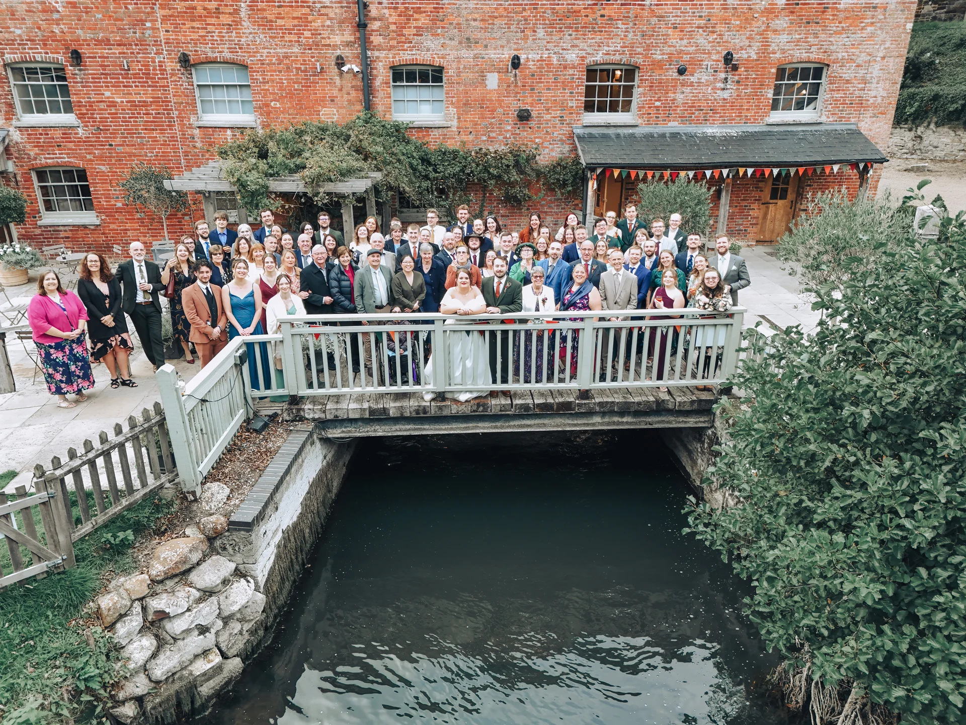 A large group of wedding guests and the newly married couple gather on a wooden footbridge over a canal in front of an exposed red brick building.