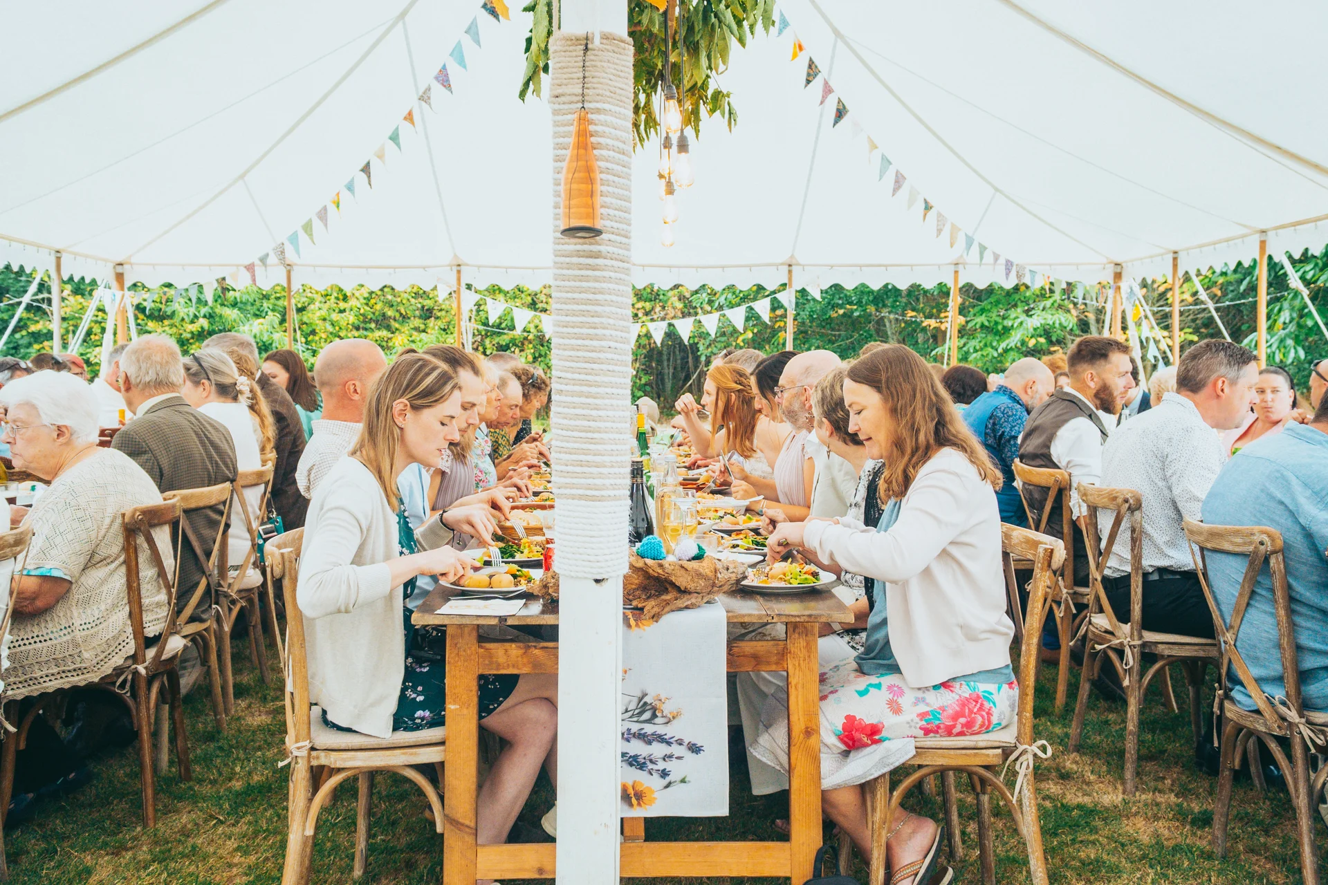 Guests enjoy a wedding breakfast seated at long wooden tables beneath a bright white marquee decorated with bunting.