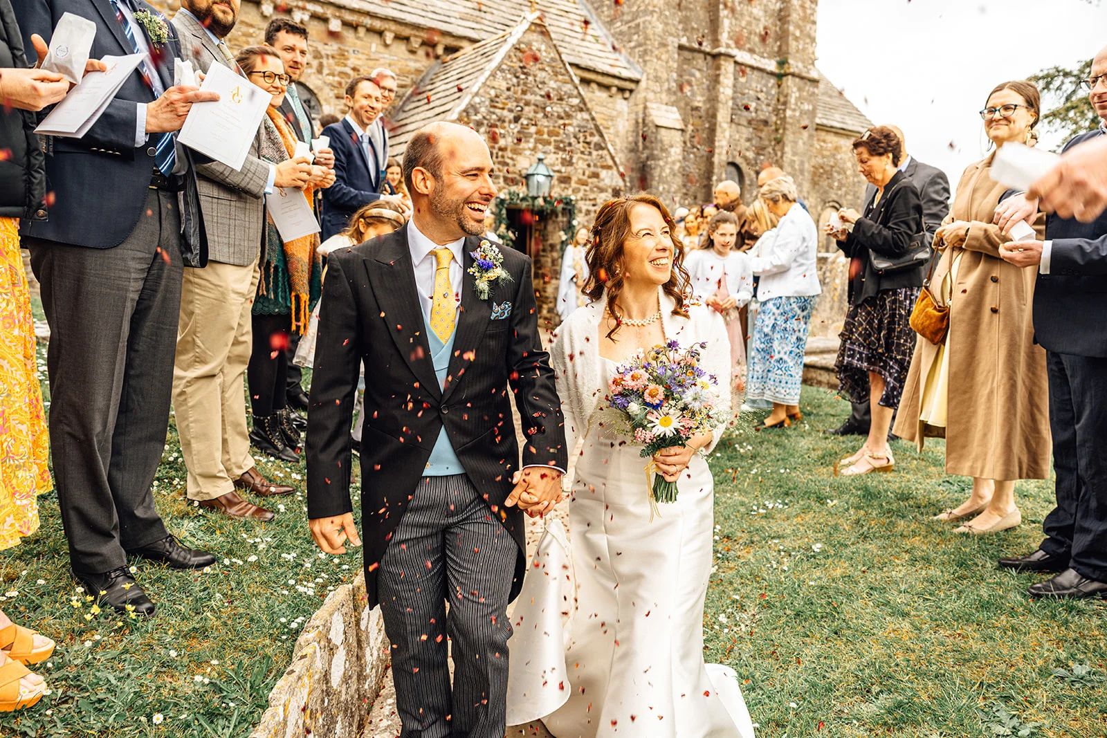 A joyful newlywed couple walks hand in hand out of a stone church while guests shower them with celebratory confetti.