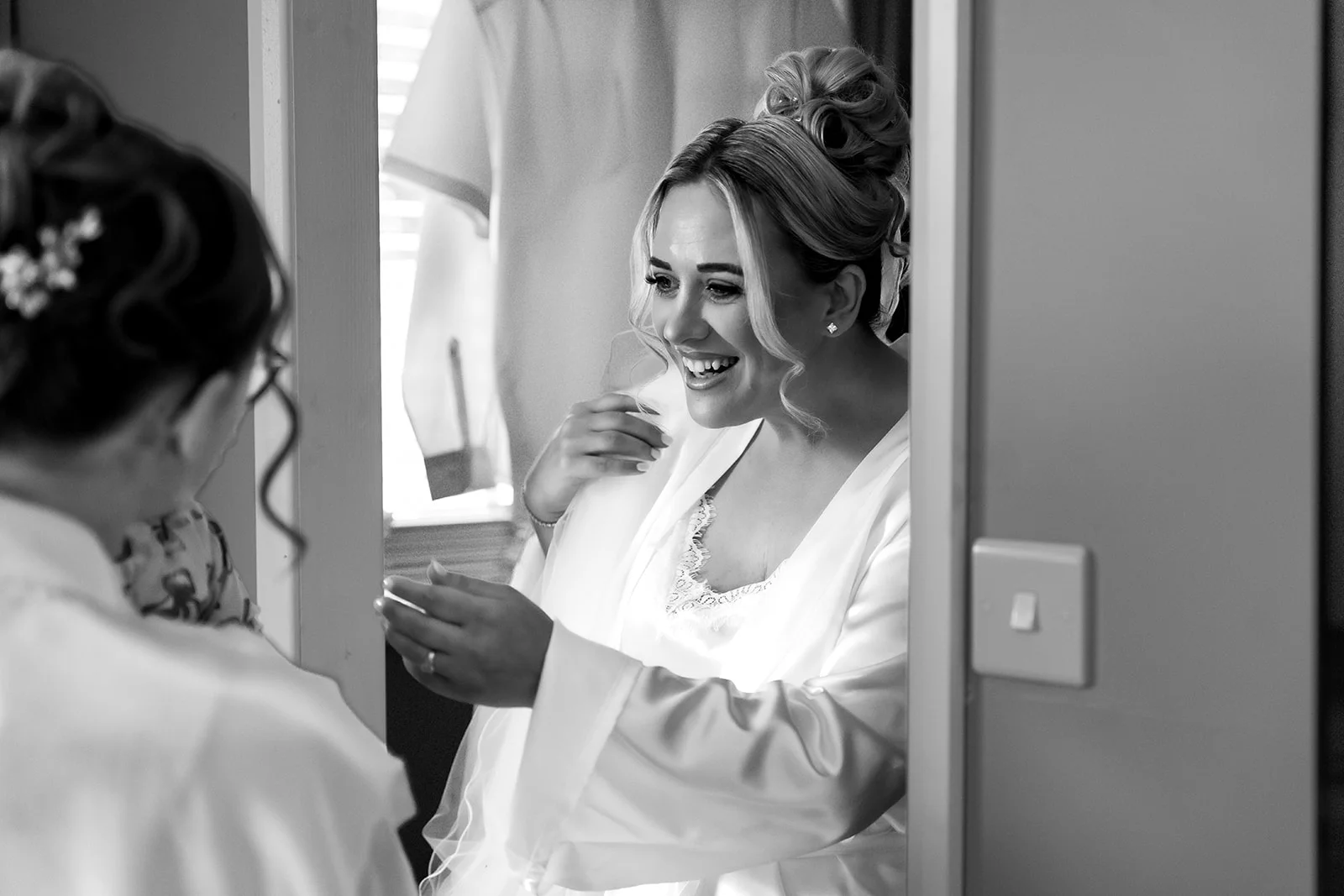 A smiling bride with her hair in a high bun wearing a satin getting ready robe laughs joyfully while looking through a doorway.
