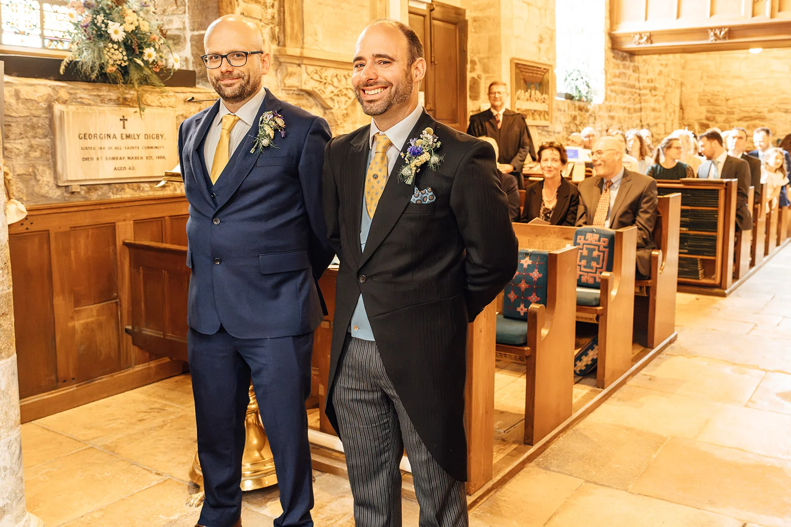 Two grooms in sharp suits stand smiling inside a historic stone church while guests are seated in wooden pews behind them.