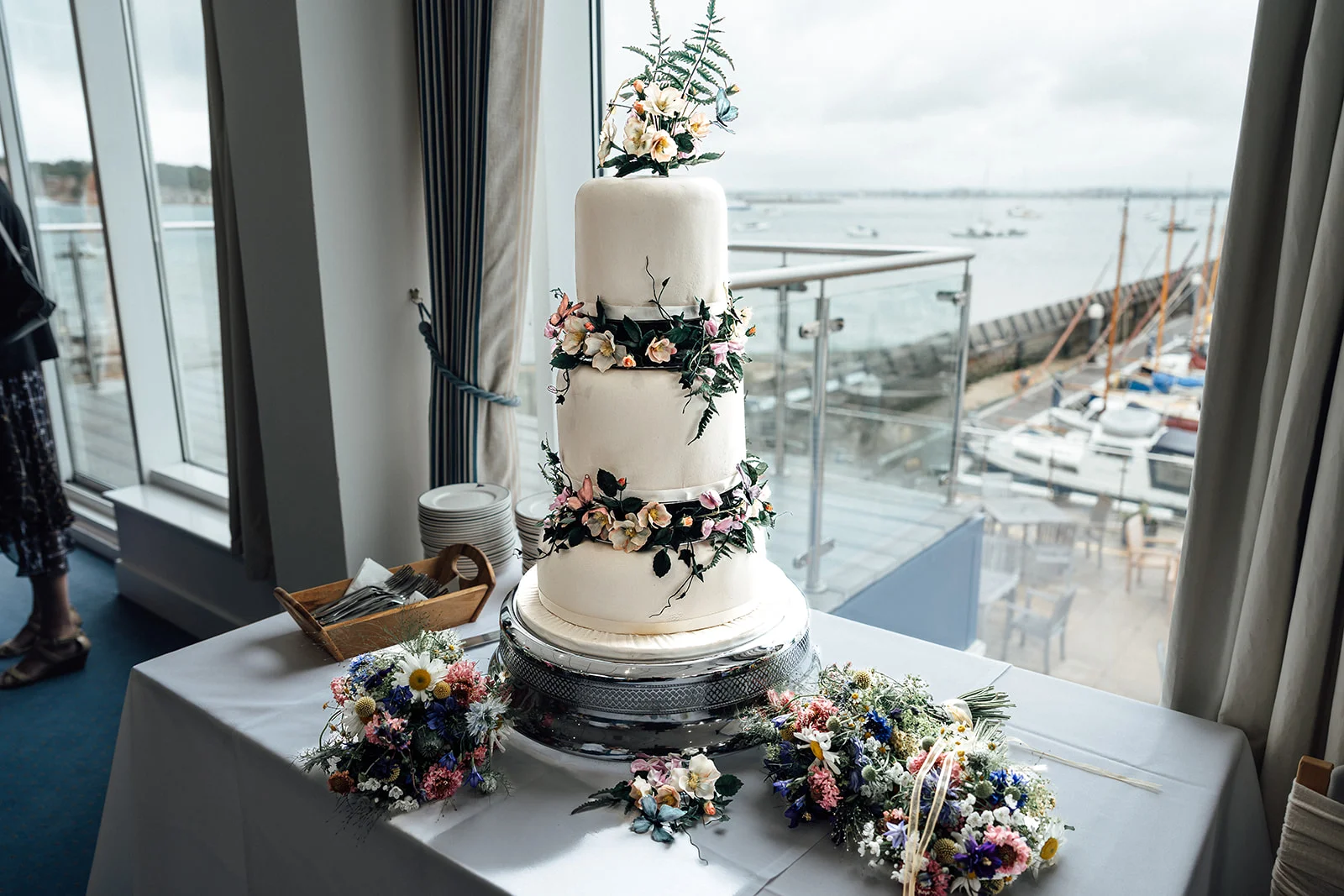 A four-tier white wedding cake adorned with delicate floral cascades stands on a table next to colourful bouquets, with a scenic harbour view visible through large windows behind.