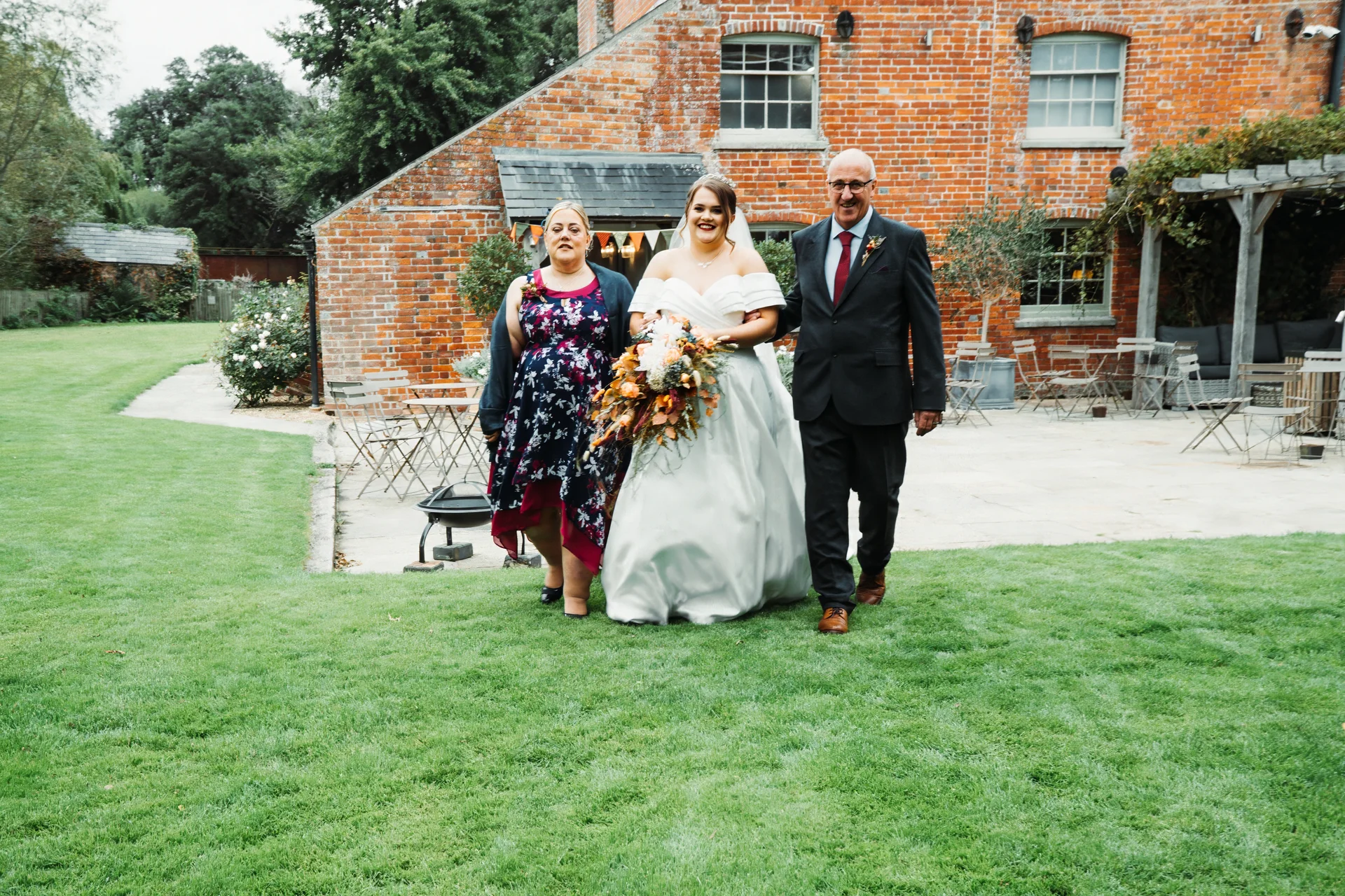 A smiling bride walks down an outdoor aisle across a lush green lawn, linked arm in arm with her parents in front of a rustic red brick venue.