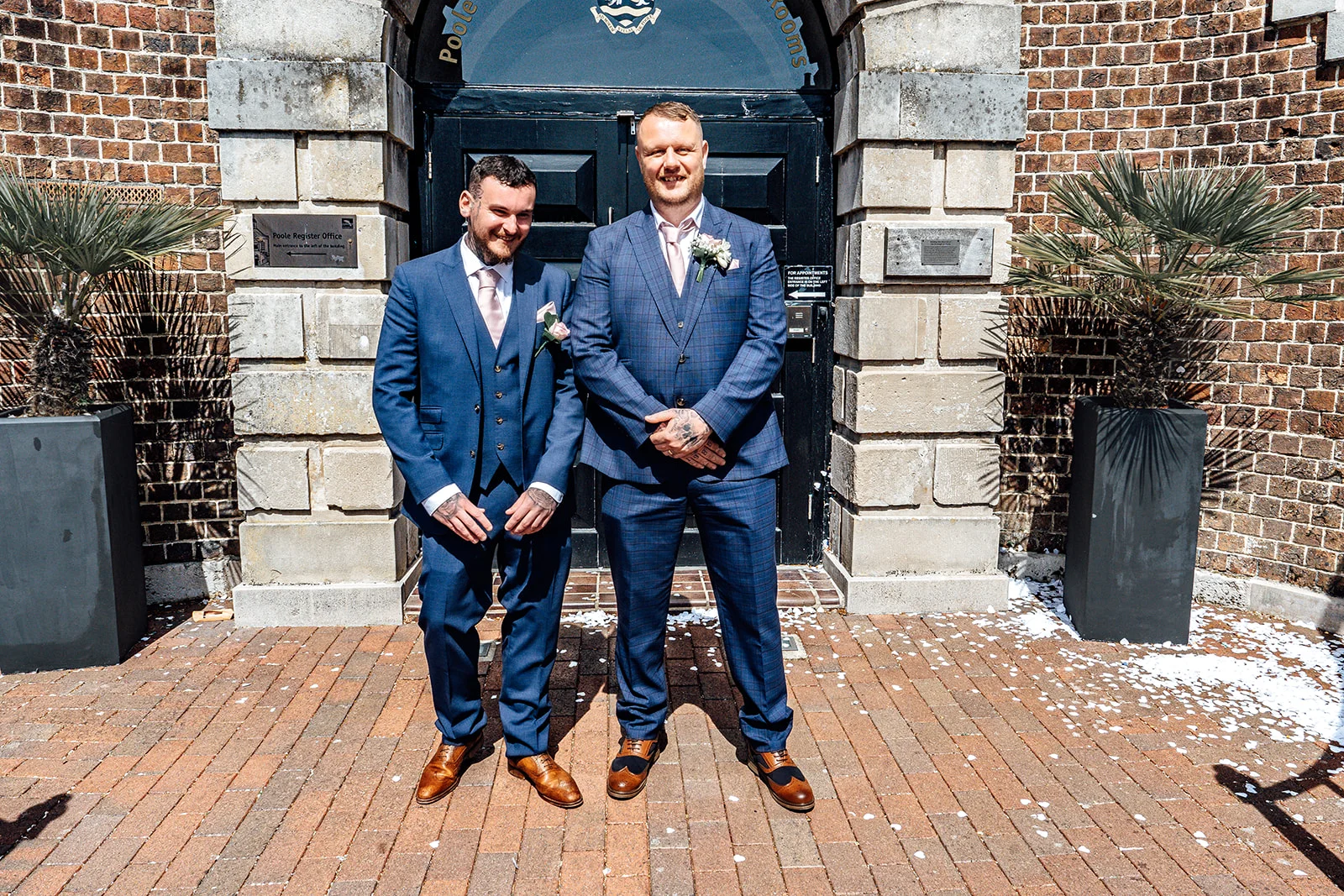 Two grooms in matching navy and checked three-piece suits stand smiling outside the brick entrance of Poole Register Office on a sunny day.