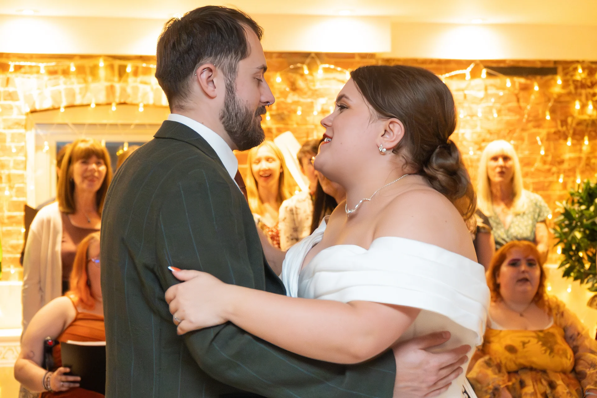 A newlywed couple shares their first dance together indoors against a warm brick backdrop illuminated by twinkling fairy lights, surrounded by smiling guests.