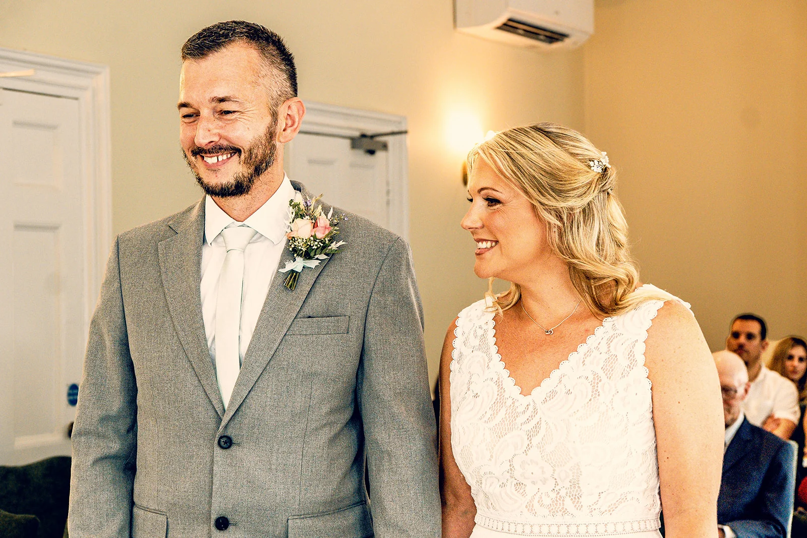 A smiling groom in a grey suit and the bride in a lace wedding dress stand together during their wedding ceremony.