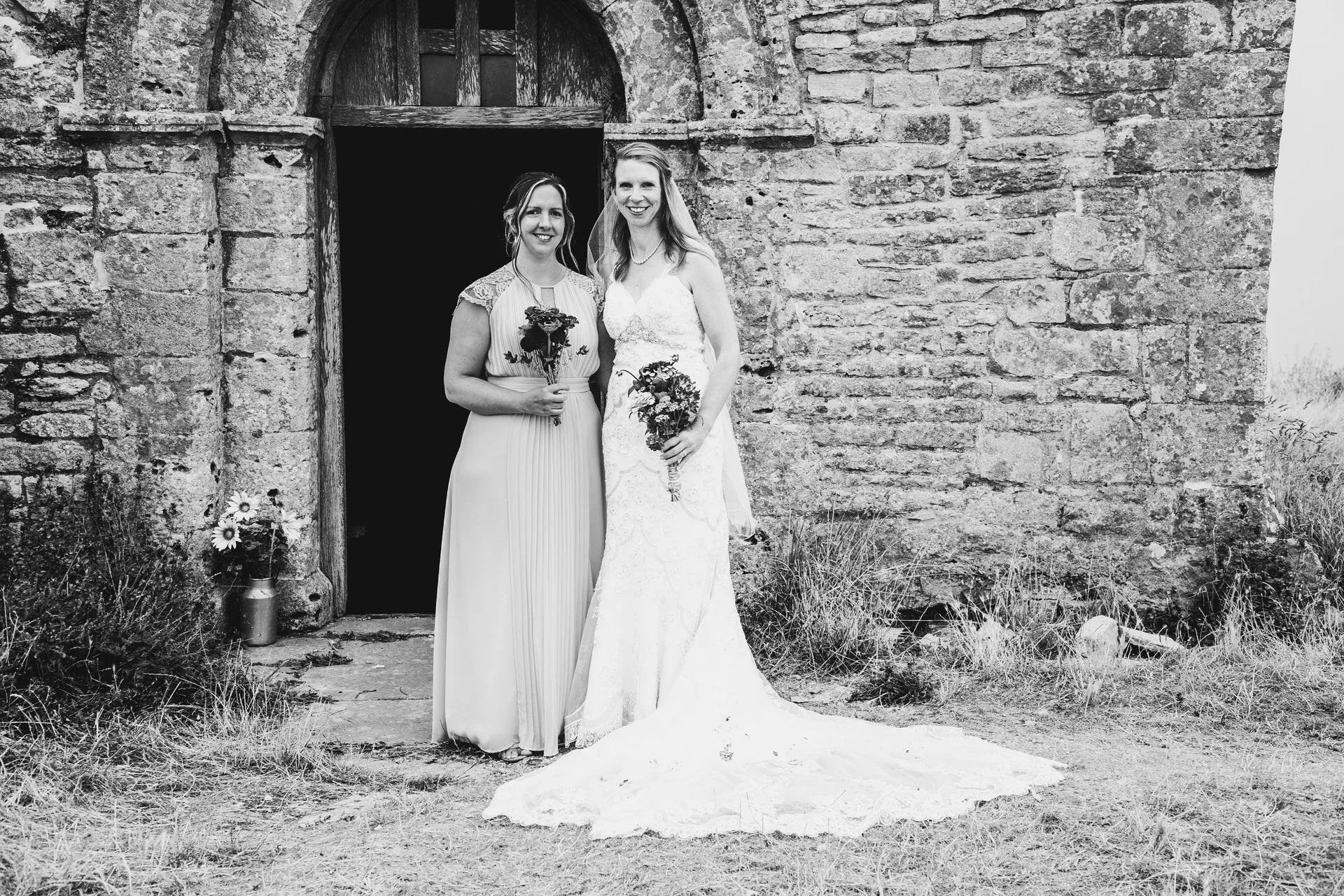 Two brides pose together outside a rustic stone church doorway holding floral bouquets in a classic black and white portrait.