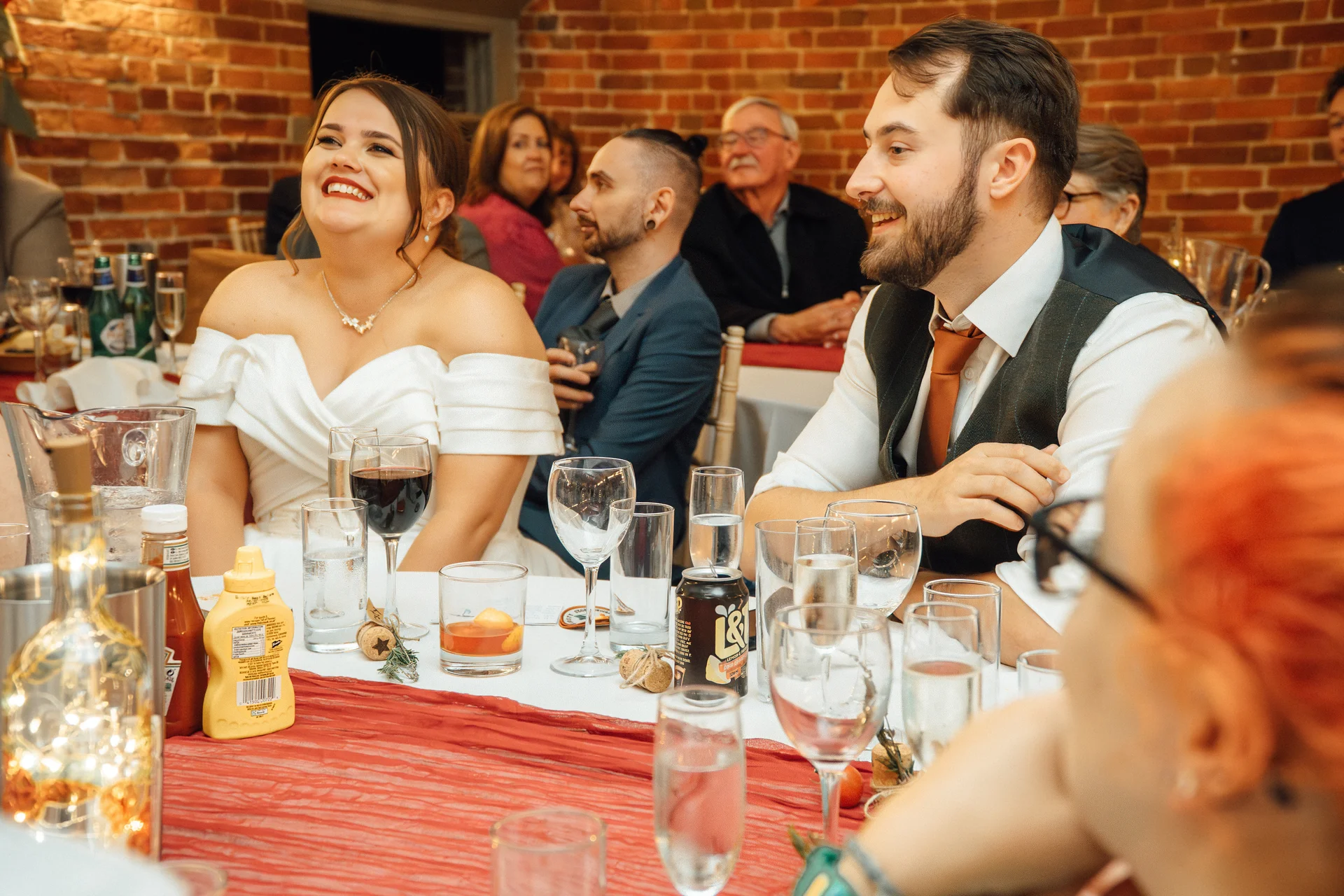 A joyful bride in an off-the-shoulder gown and the smiling groom in a waistcoat share a happy laugh while seated at the head table during the wedding reception.