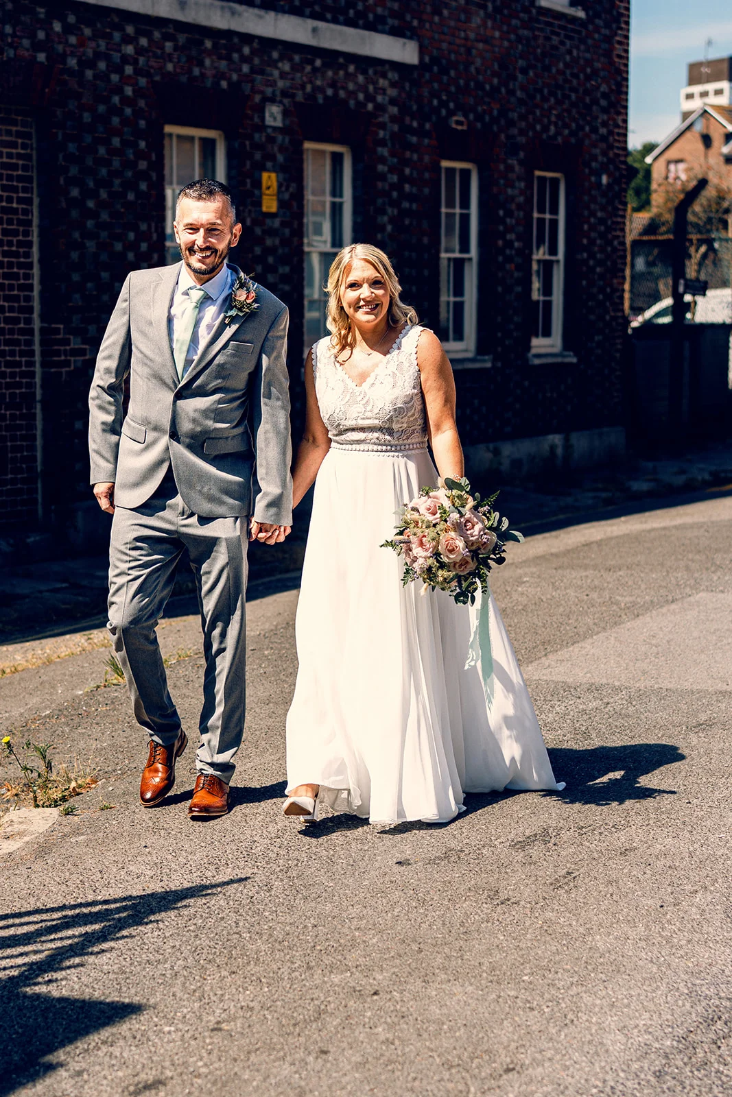 A smiling bride and groom holding hands and walking together along an urban street in bright sunshine.