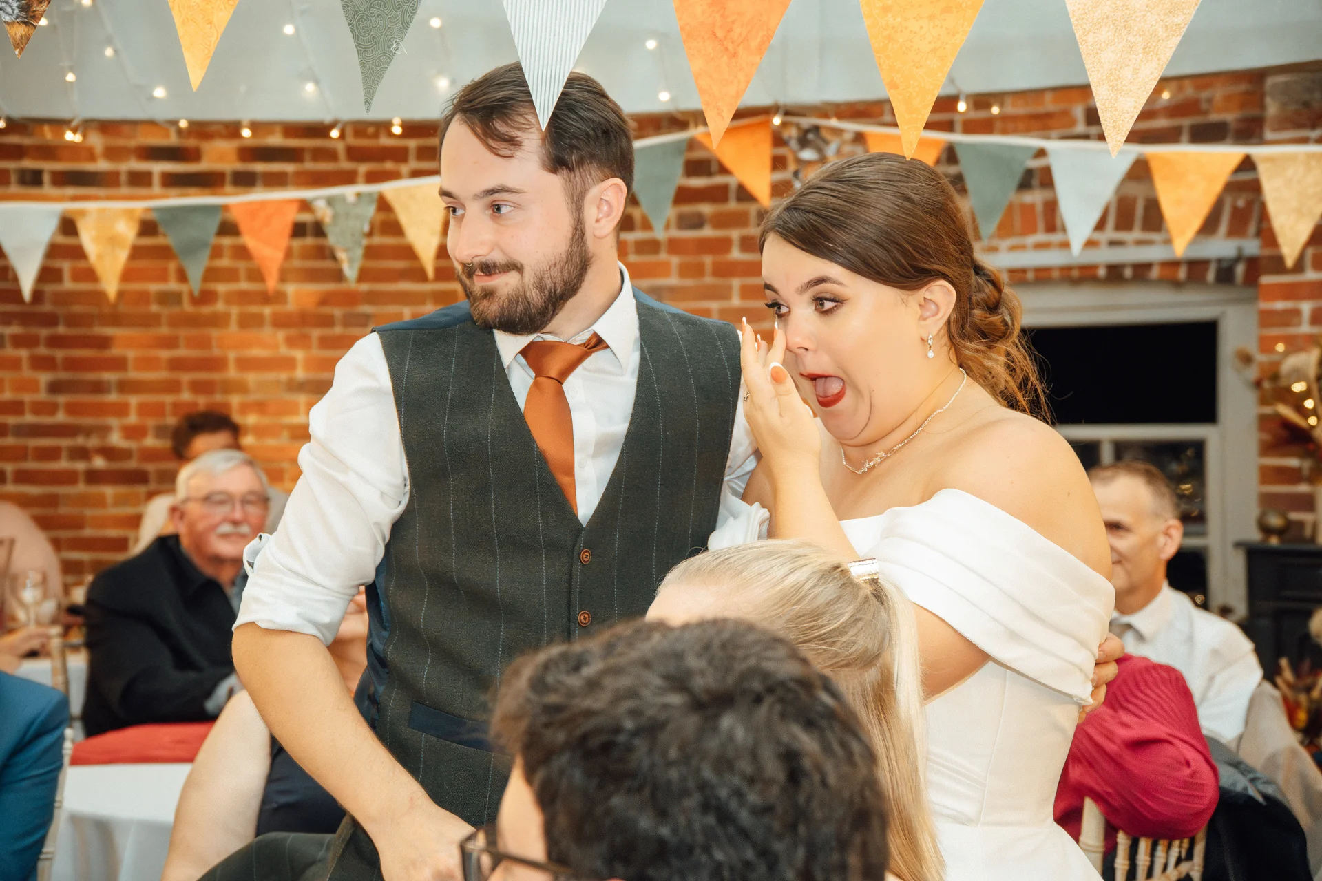 A bride reacts with shock and laughter during the wedding speeches as her groom looks on beside her in front of a rustic brick wall.