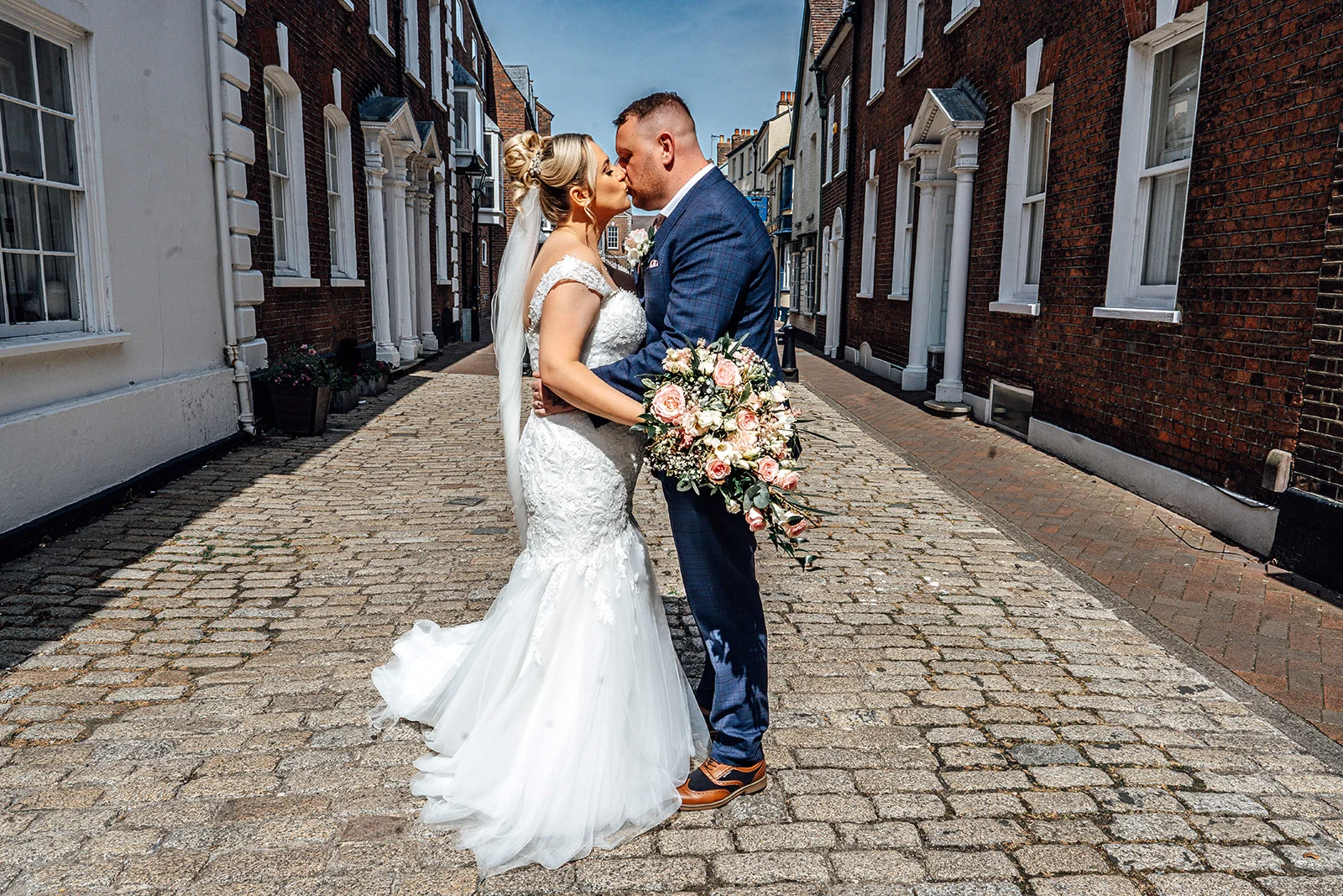 Newlywed bride and groom share a tender kiss while standing on a historic cobbled street lined with traditional brick buildings.