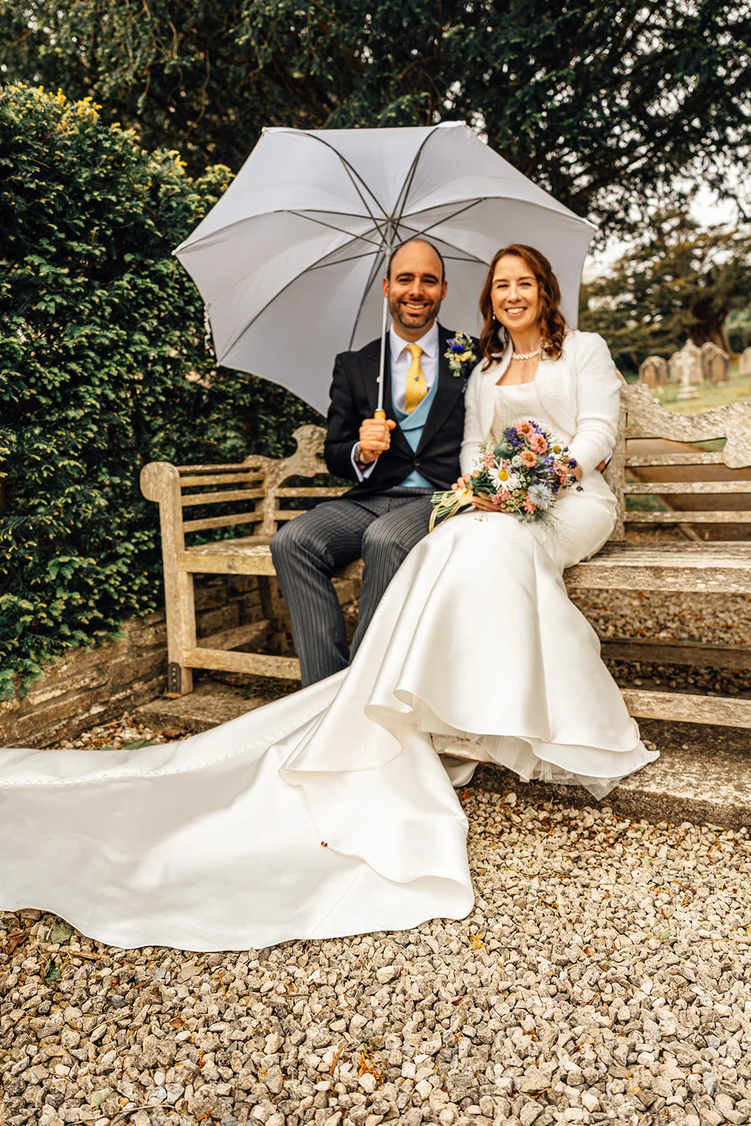 A newlywed couple smiling together on a wooden garden bench while holding a large clear umbrella on their rainy wedding day.