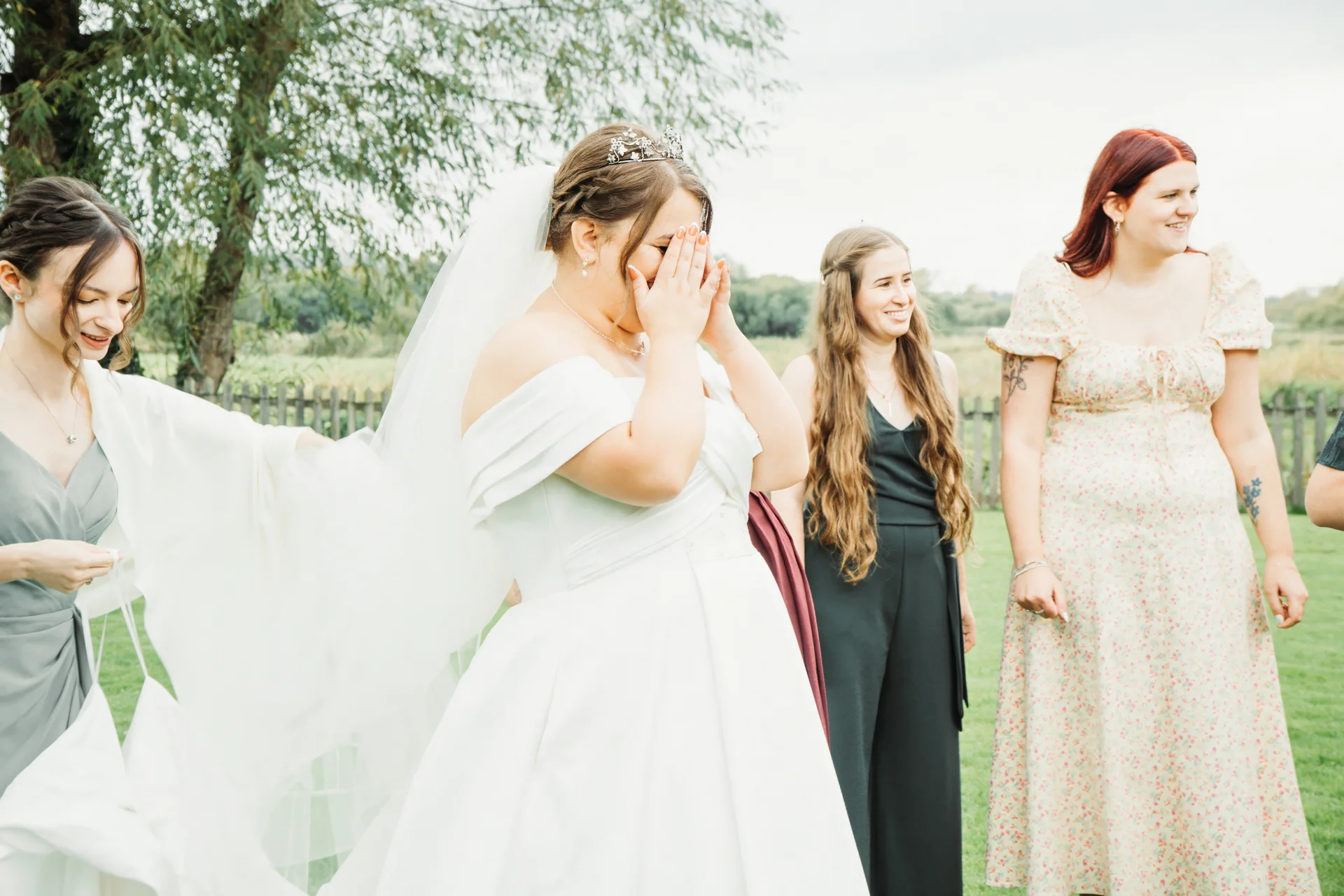 An emotional bride in an off-the-shoulder satin gown and tiara covers her face in surprise as her bridesmaids react happily outdoors.