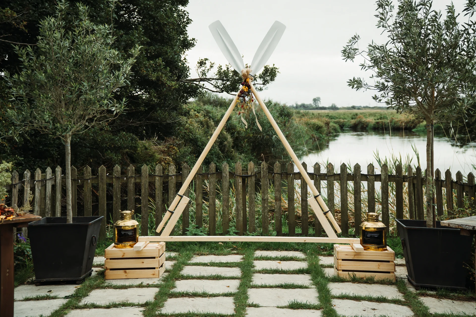 A rustic wooden triangular ceremony arch stands outdoors on a stone patio overlooking a tranquil river, flanked by potted olive trees and brass lanterns.