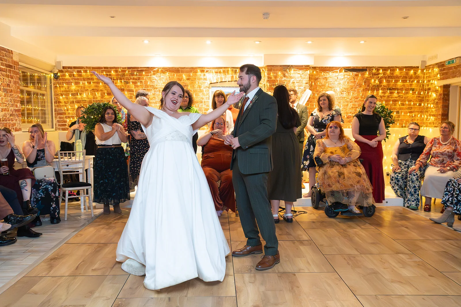 A joyful plus-size bride in an off-the-shoulder gown dances enthusiastically with her groom in a green suit, surrounded by smiling guests in a brick-walled venue illuminated by fairy lights.
