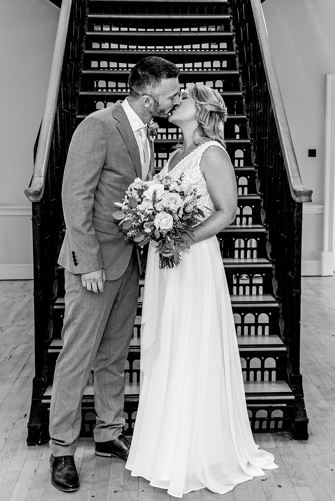 The bride and groom share a tender kiss while standing on a sweeping staircase holding a floral bouquet.
