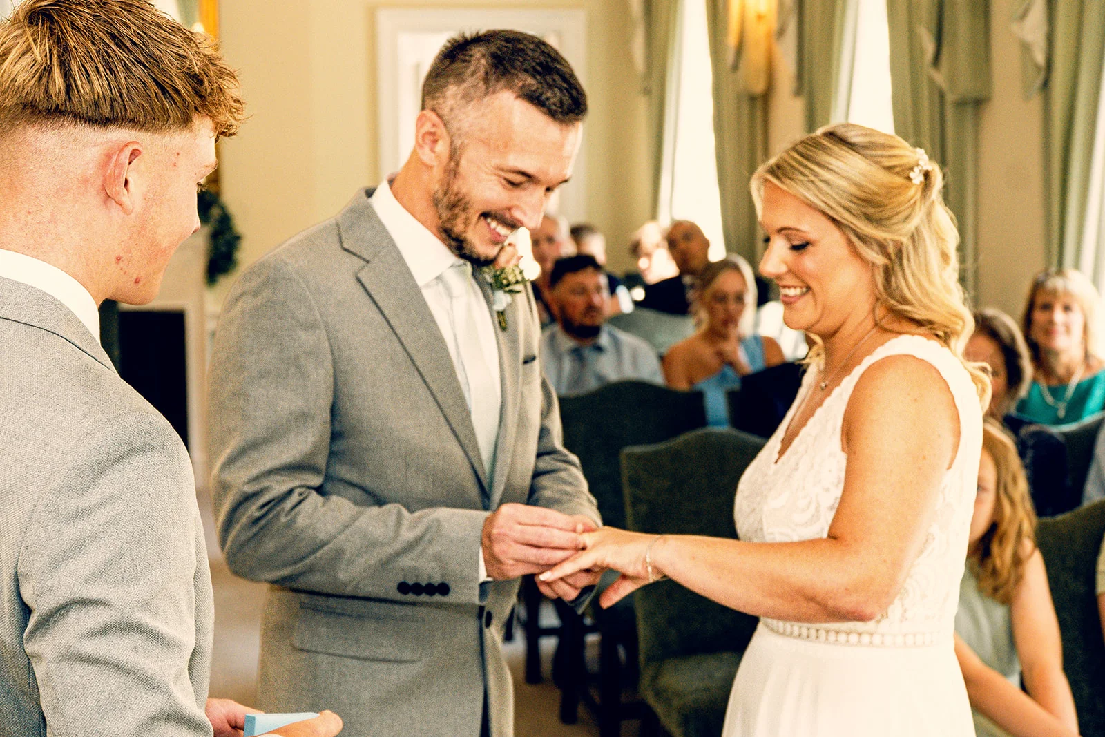 A smiling groom places a wedding ring onto the bride's finger during their indoor ceremony as guests watch from seated rows.