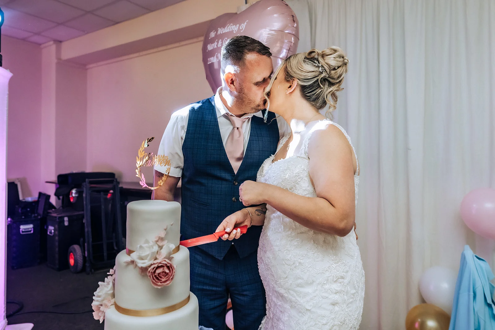 Newlyweds share a tender kiss while cutting their elegant three-tiered white wedding cake adorned with pink floral accents.