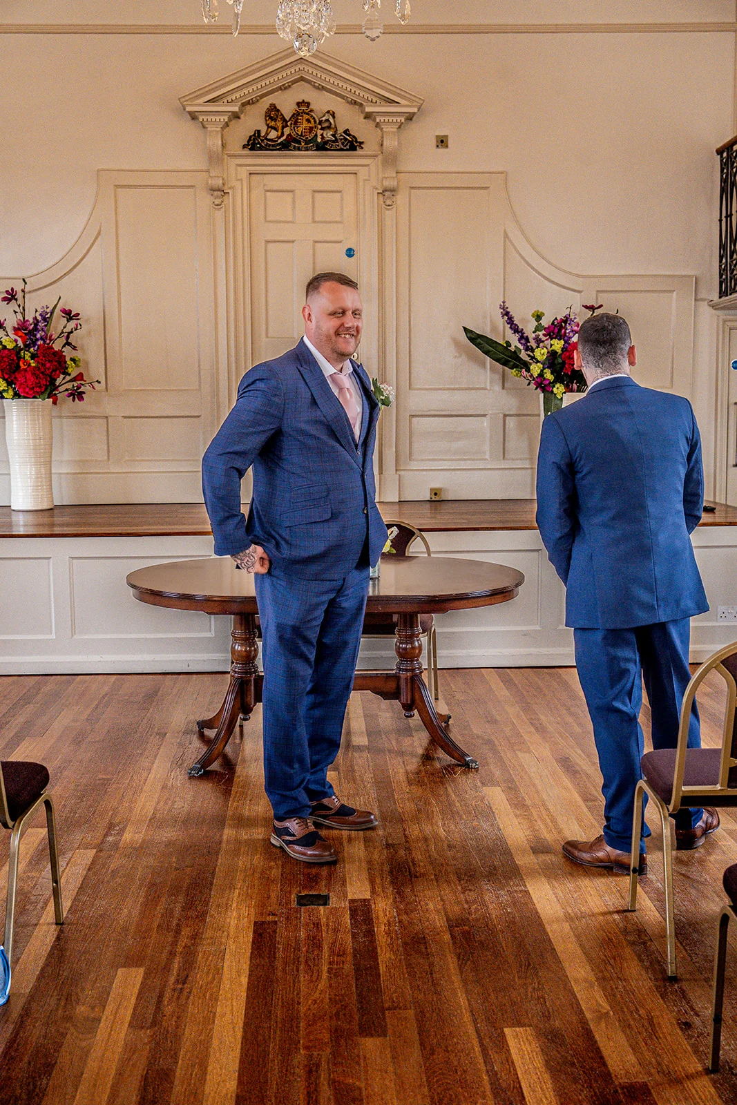 Two grooms in matching blue suits share a joyful first look moment inside an elegant wood-panelled registry office.