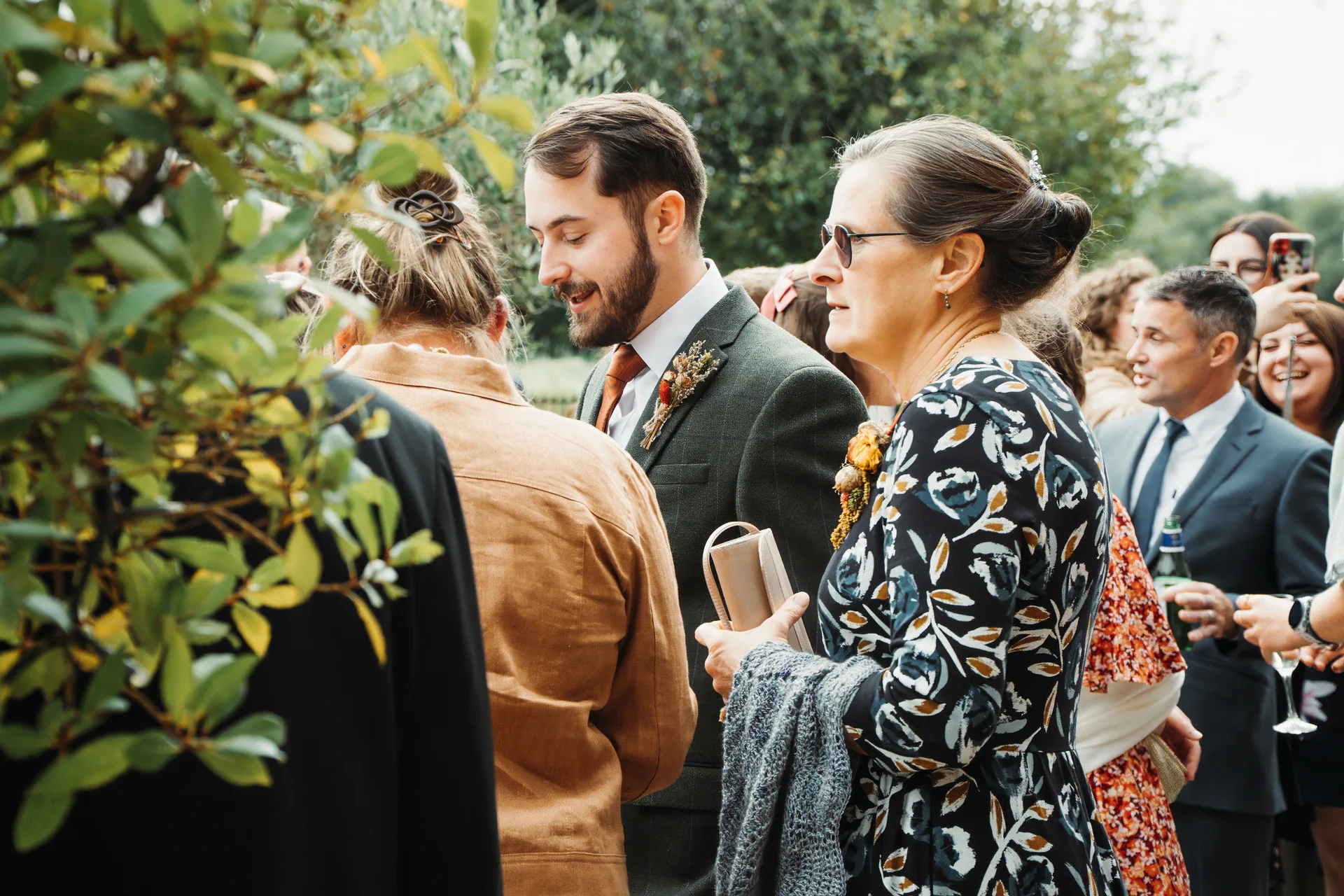 Wedding guests mingle outdoors in a sunlit garden, with a man in a green tweed suit and a woman in a floral dress conversing.