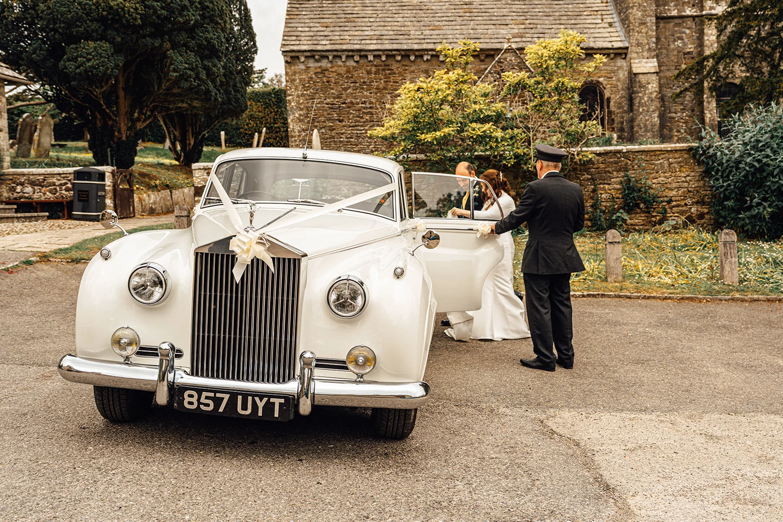 A chauffeur in uniform opens the door of a classic white vintage wedding car outside a historic stone church.