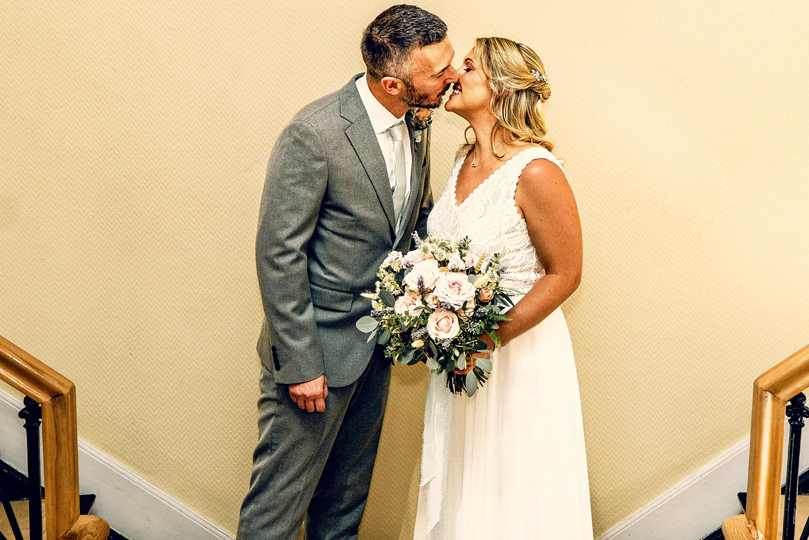 A newlywed couple shares a tender kiss indoors near a staircase, with the bride holding a delicate floral bouquet.